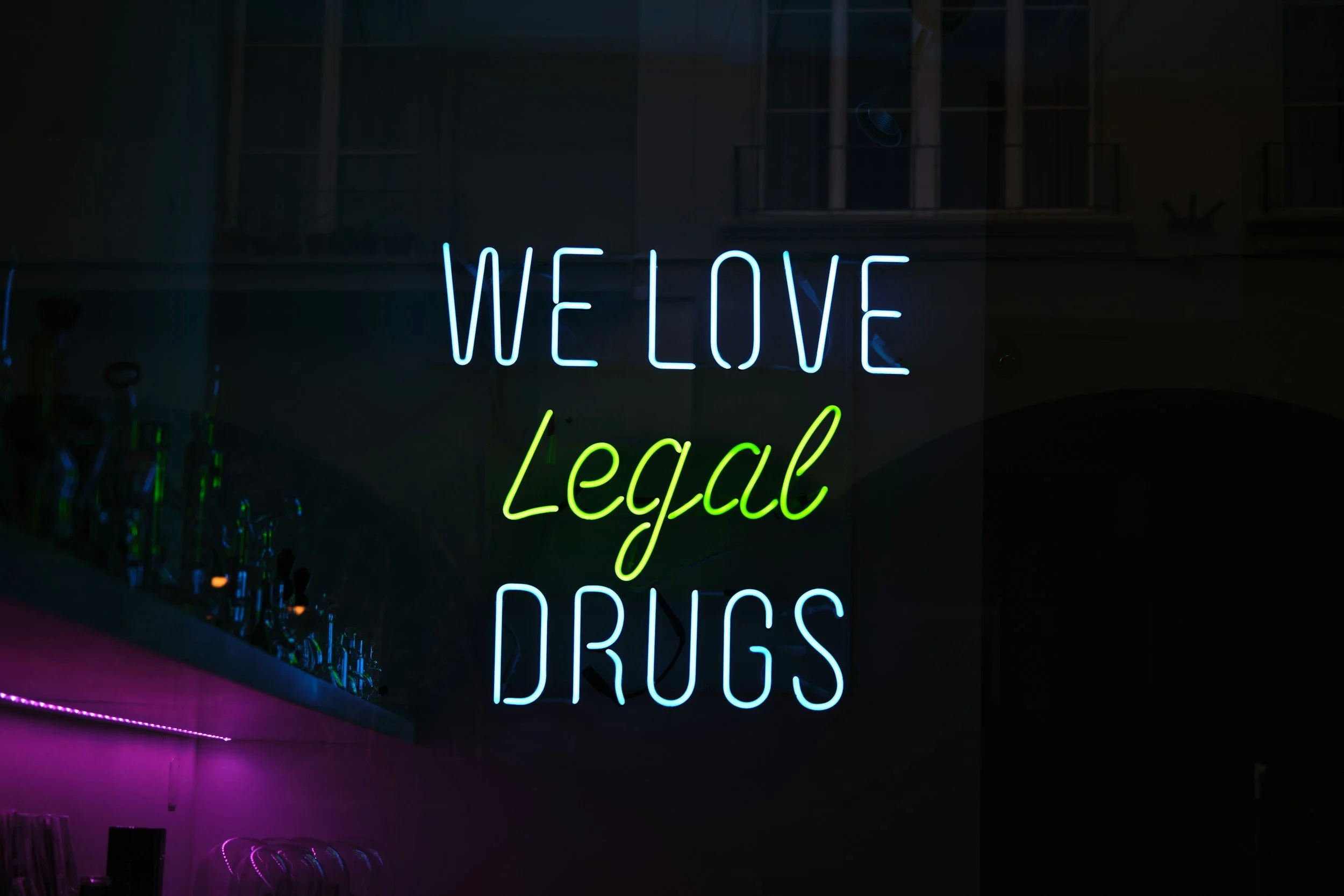 A neon sign with the words "WE LOVE Legal DRUGS" in blue, green, and pink colors.