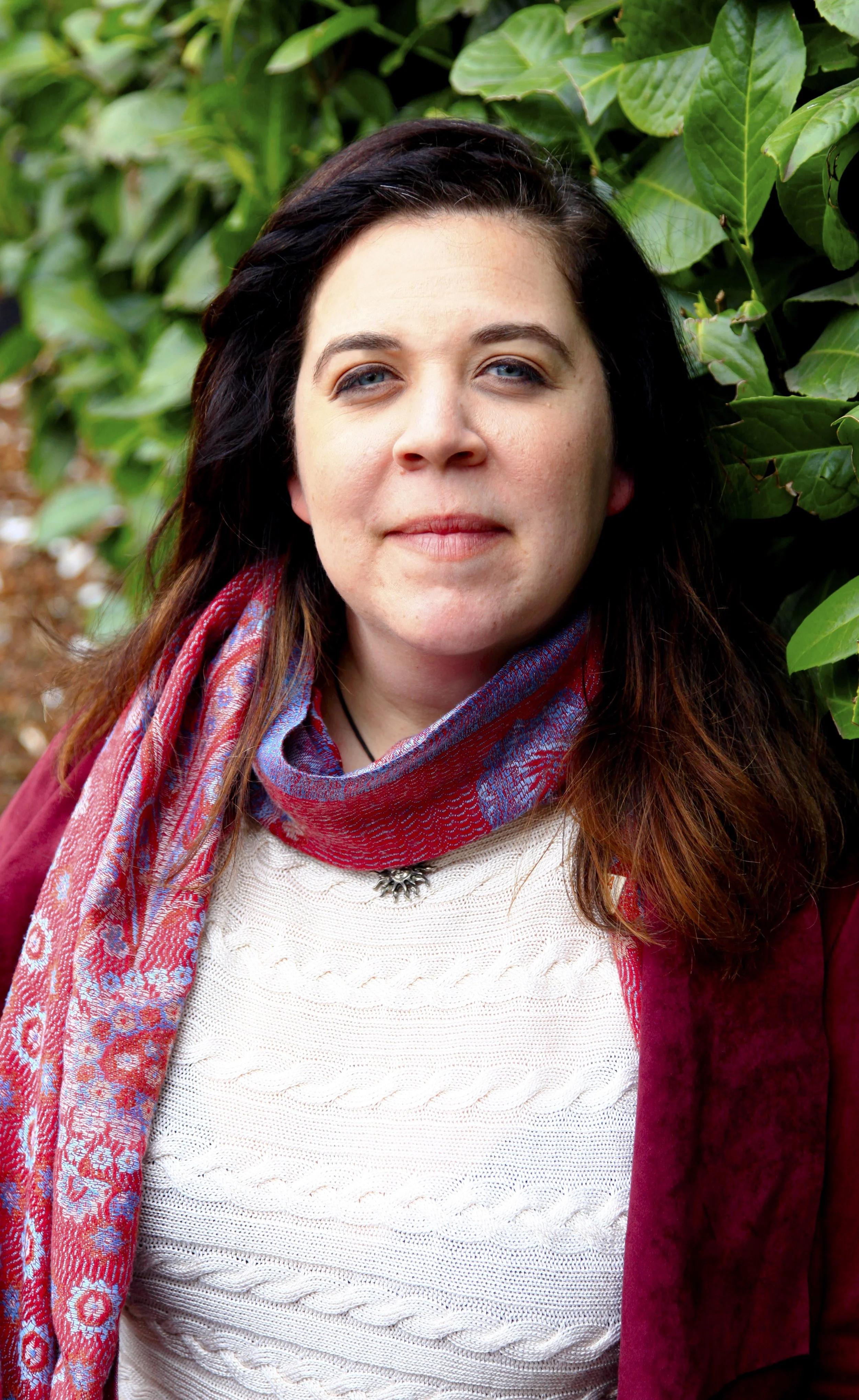 A woman with dark brown hair and blue eyes standing outdoors near green leafy plants, wearing a white knitted sweater, burgundy jacket, and a patterned scarf.