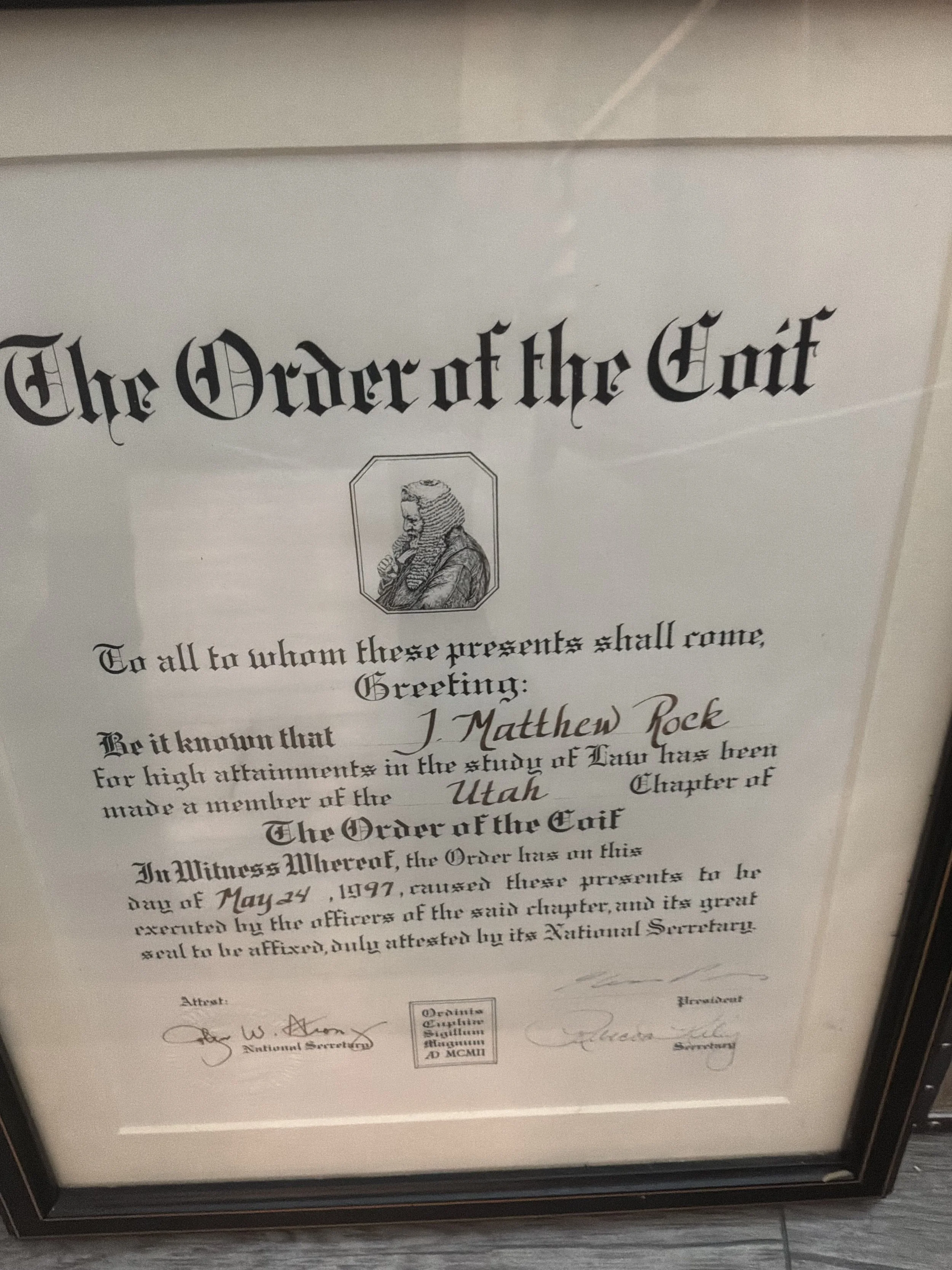 A framed document titled 'The Order of the Coif' with a black and white illustration of a judge or lawyer at the top center, and signatures at the bottom, resting on a wooden surface.