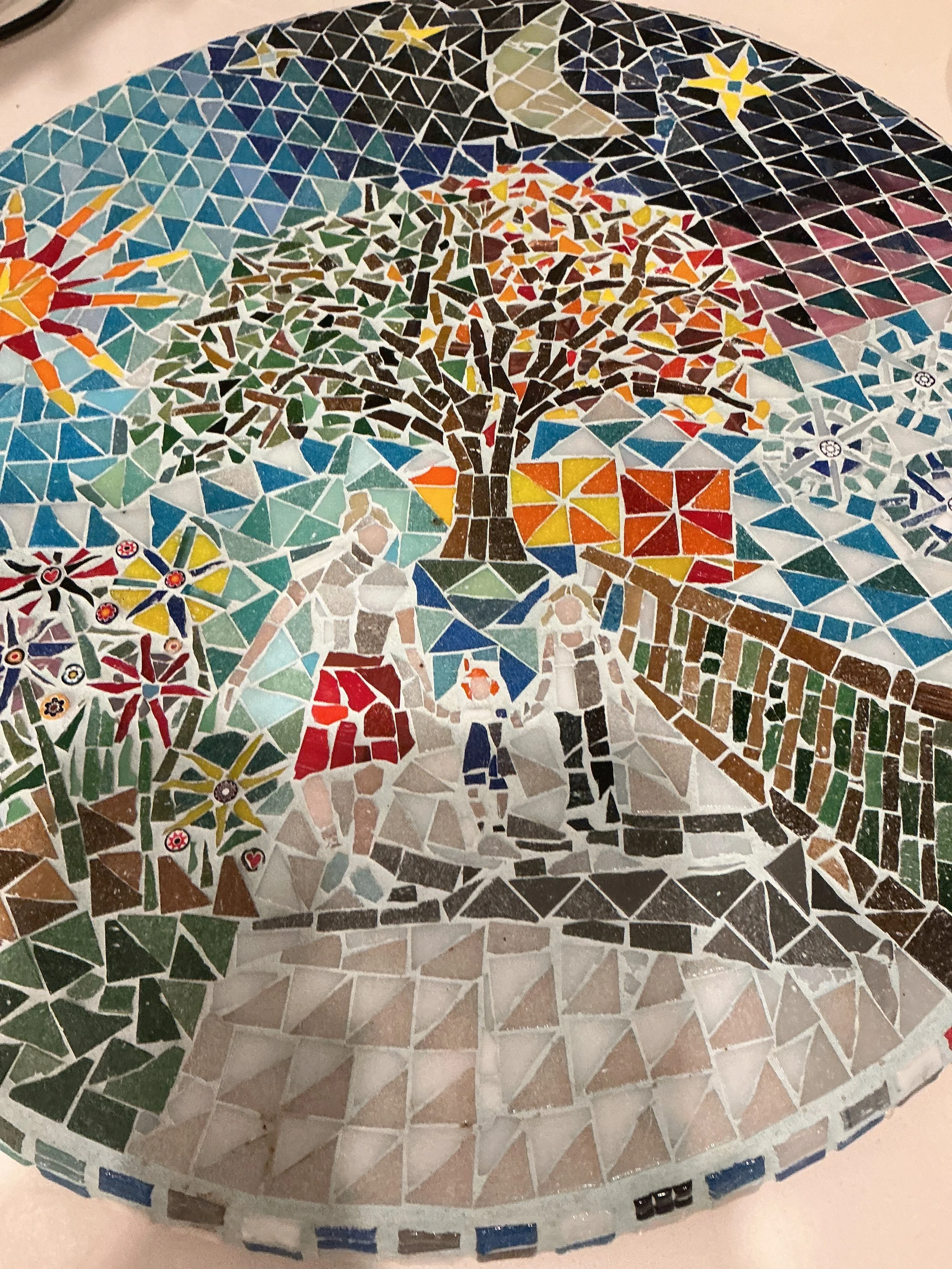 Colorful mosaic artwork depicting a family walking on a path in a park, with a large tree, flowers, a fence, a sunrise, and a moon in the background.