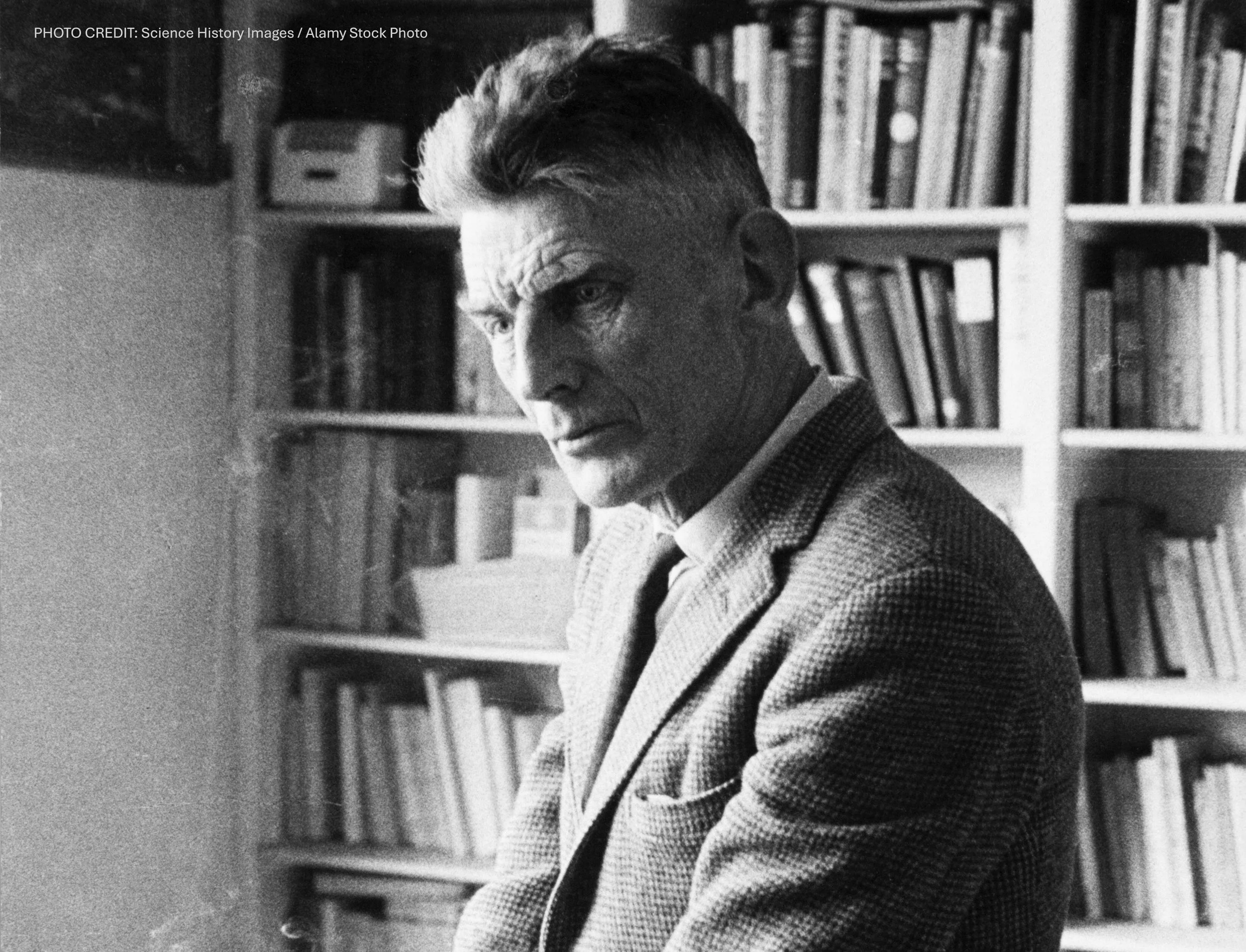 Black and white photo of a man with a serious expression, dressed in a blazer, sitting indoors with bookshelves filled with books in the background.