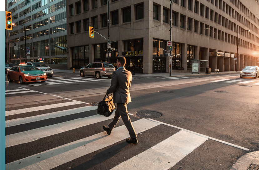 A man in a suit crossing a city street at sunset with cars passing by and tall office buildings in the background.