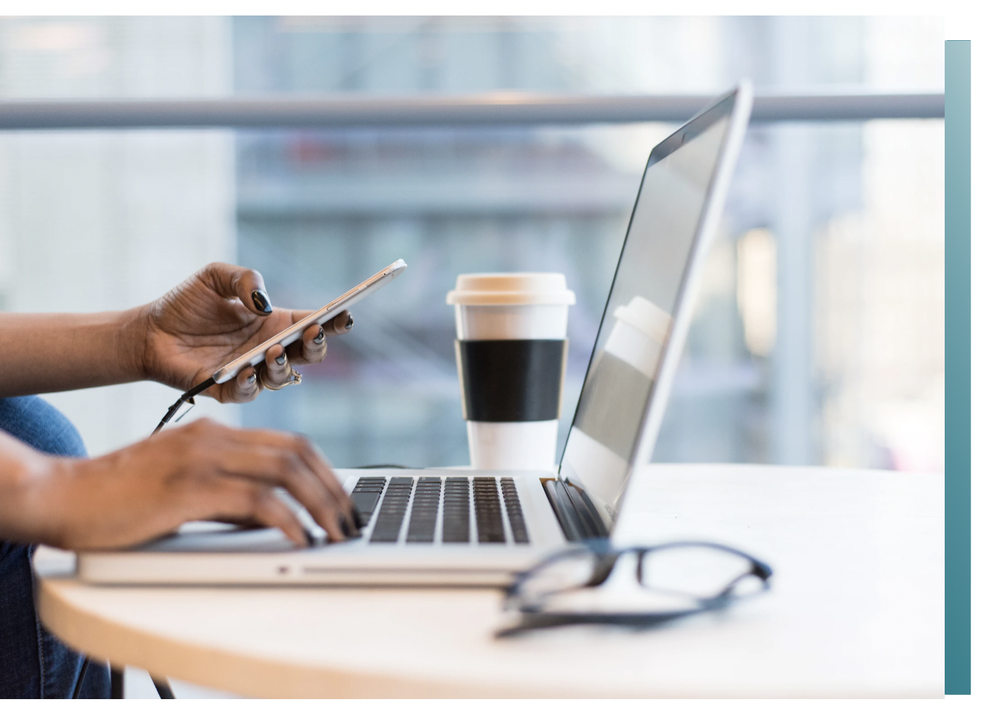Person using smartphone and working on laptop at a table with a coffee cup and glasses