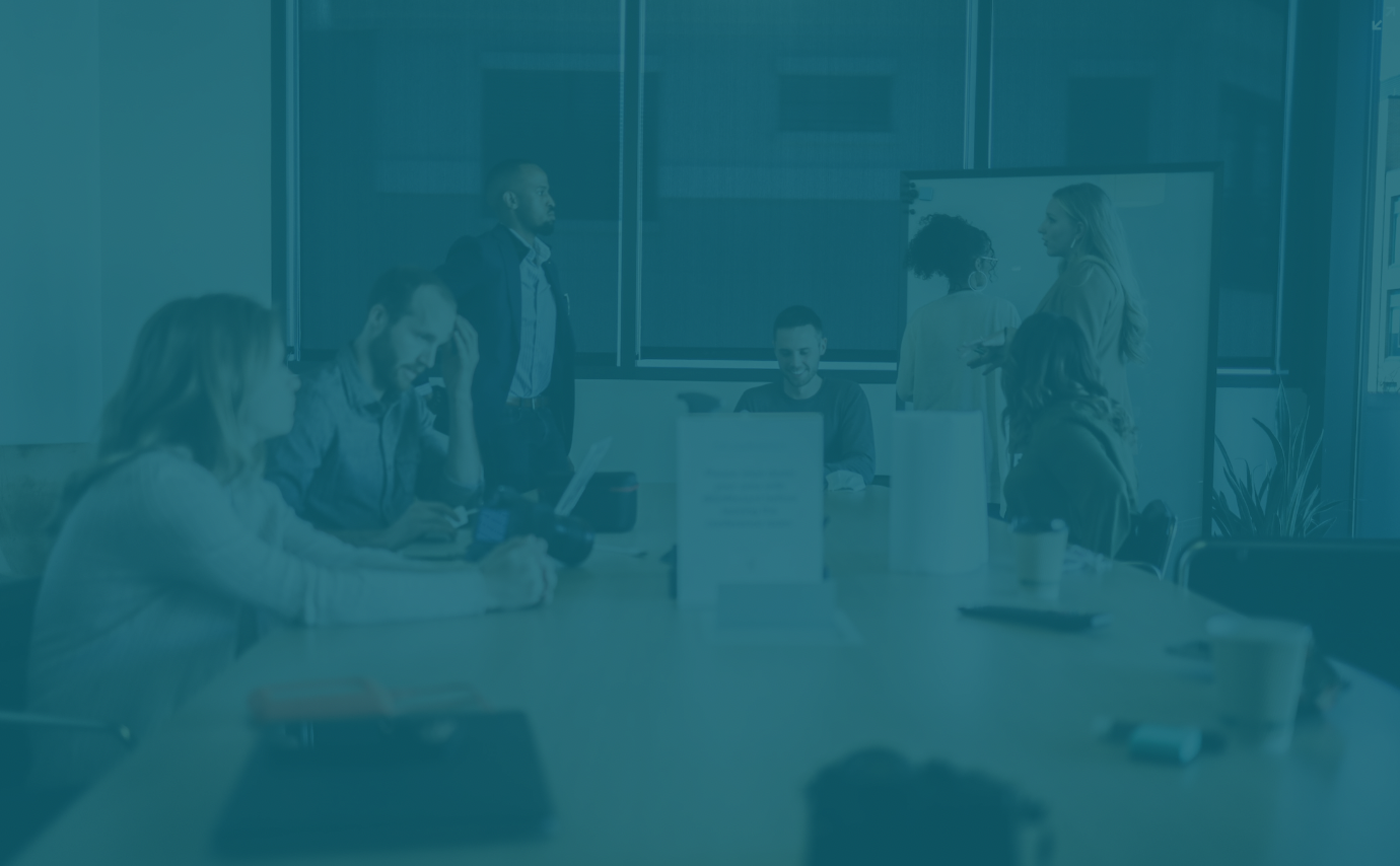 Business meeting with six people in a conference room, some are seated at a table with laptops and phones, others are standing and talking near a whiteboard.