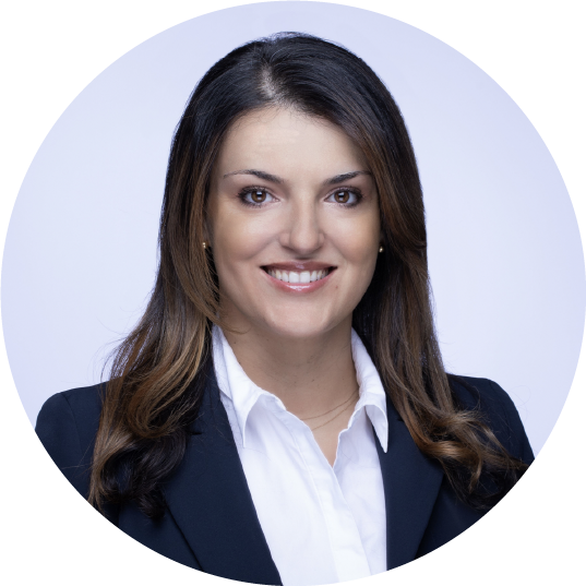 Professional woman with long brown hair smiling, wearing a white shirt and a dark blazer, against a light background.