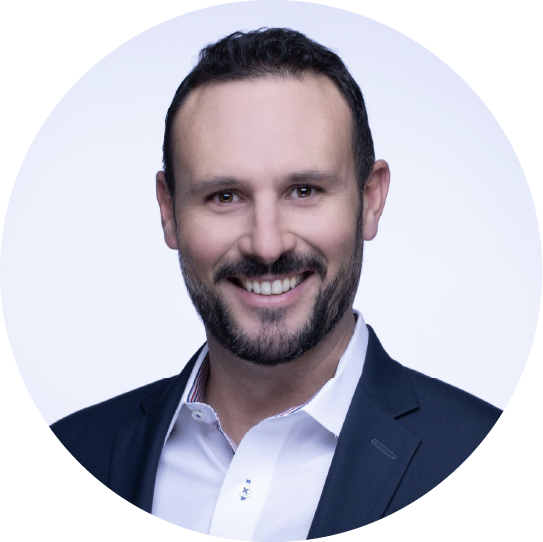 A smiling man with dark hair, a beard, and a mustache, wearing a white shirt and a dark blazer, looking at the camera against a white background.
