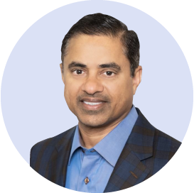 A man with short dark hair, wearing a blue shirt and a dark plaid blazer, smiling against a light blue background.