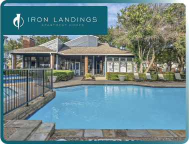 View of a swimming pool in front of a clubhouse at Iron Landings Apartment Homes with outdoor seating and lush trees.