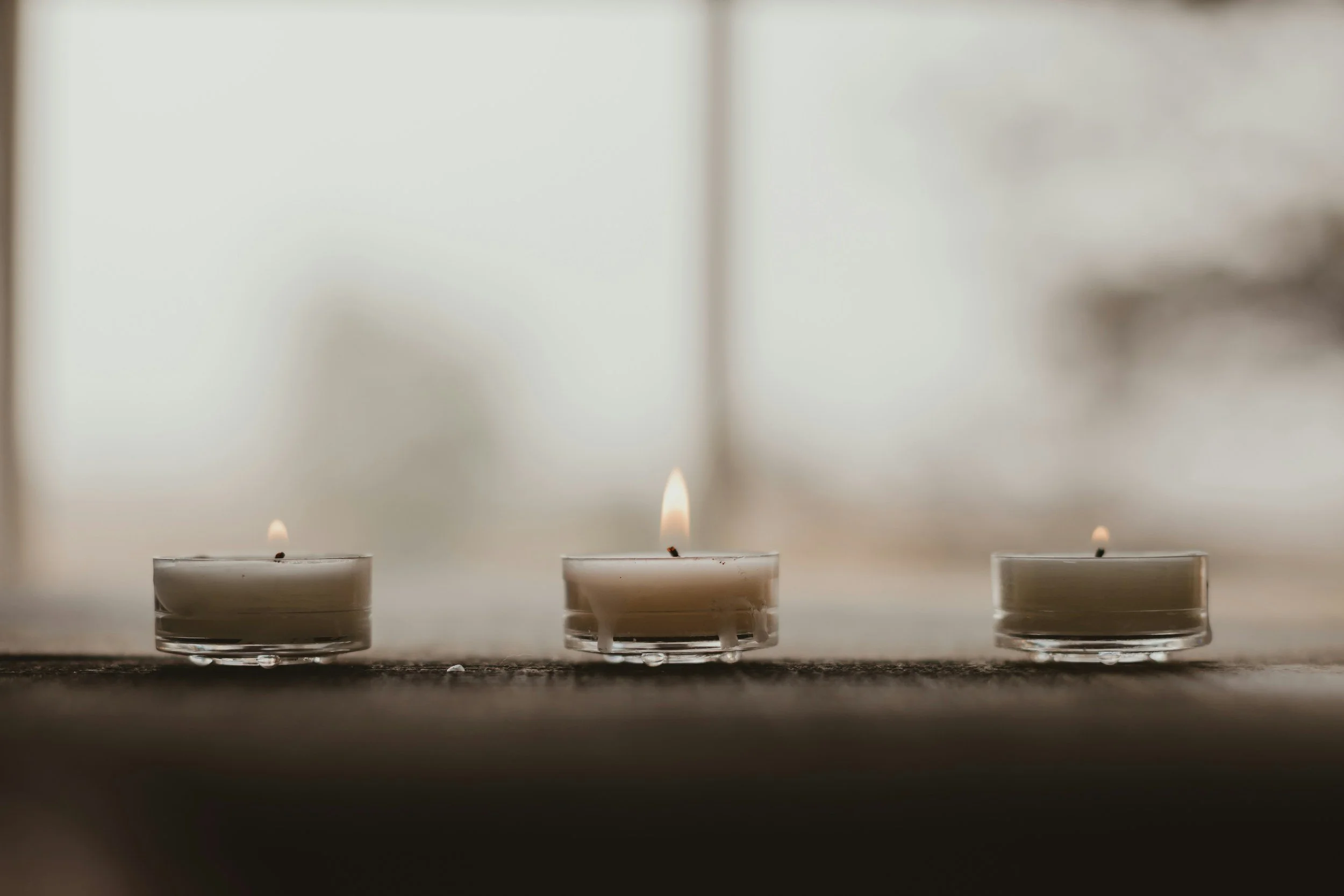 Three floating candles with lit wicks, positioned on a surface near a window, with a blurry background.