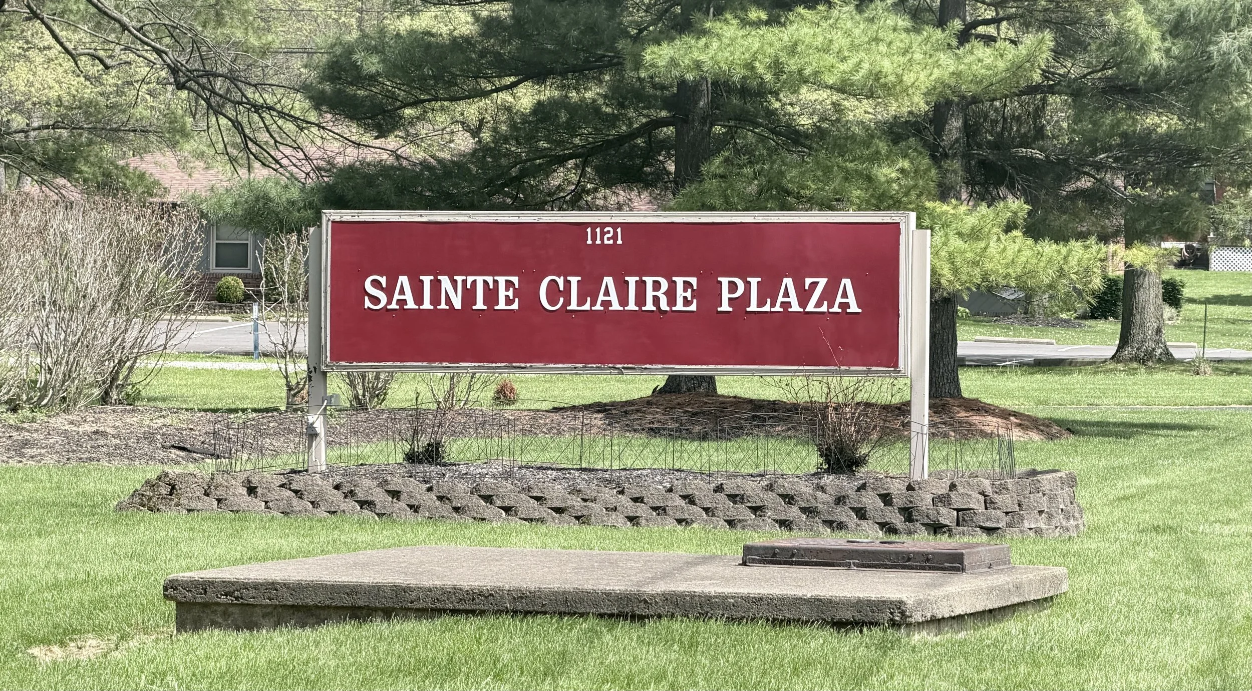 A red sign with white text reading '1121 SAINTE CLAIRE PLAZA' located on a grassy area with trees and shrubs in the background.