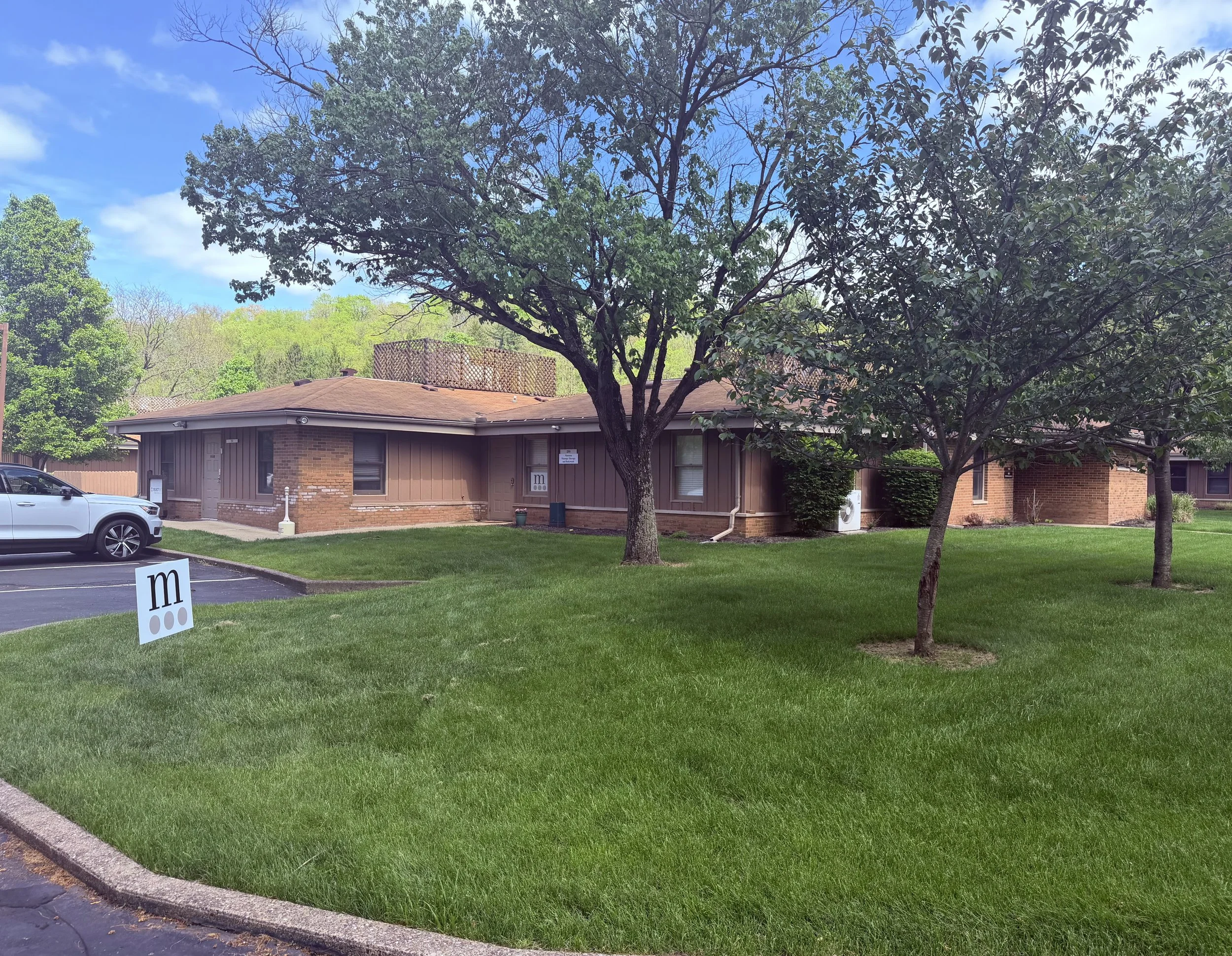 A residential building with a brown exterior and a roof with solar panels, surrounded by green trees and a manicured lawn, under a partly cloudy blue sky.