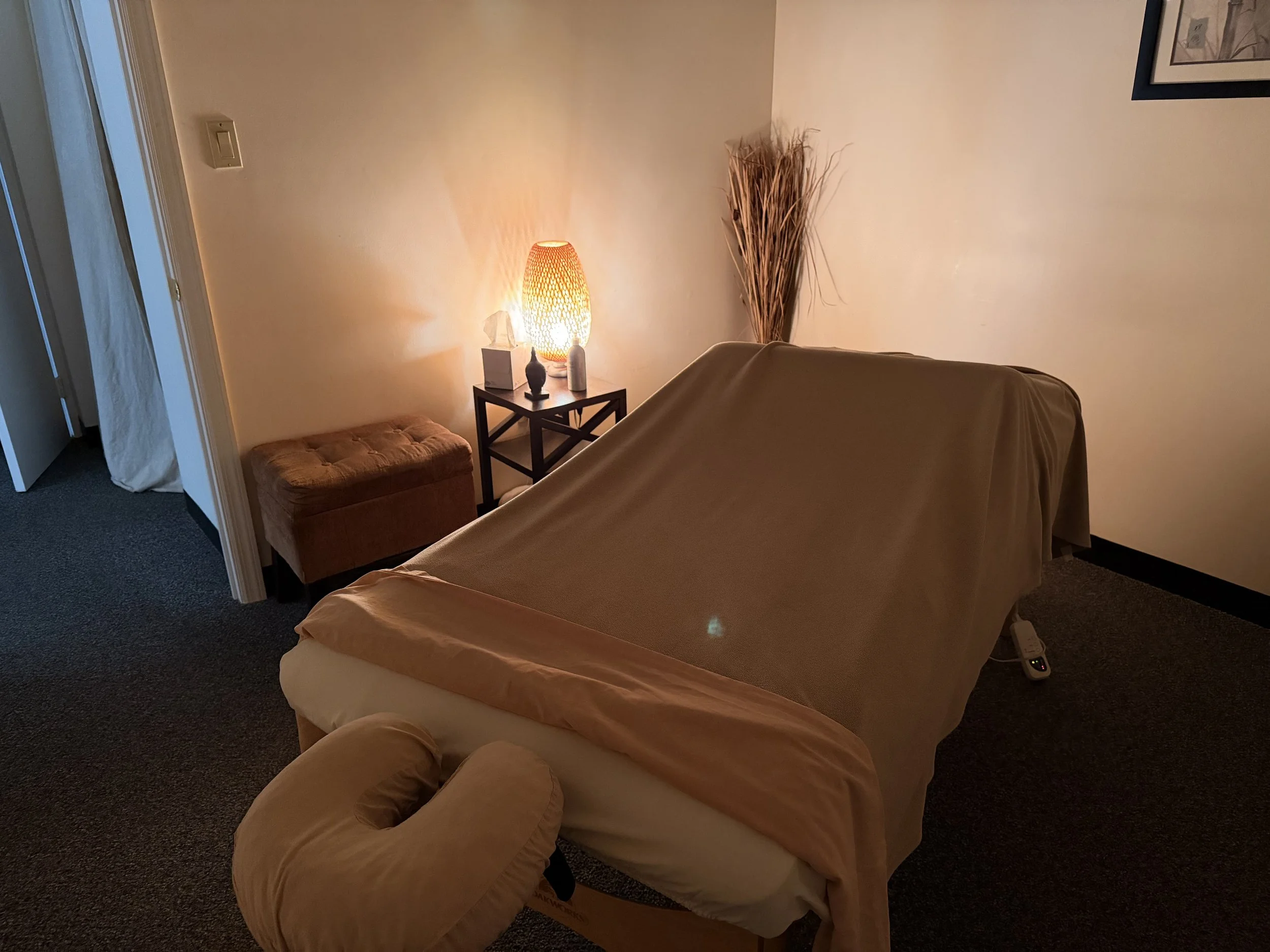 A massage table covered with a beige sheet in a dimly lit room with a lamp on a small black table, decorative objects, and a tufted ottoman.