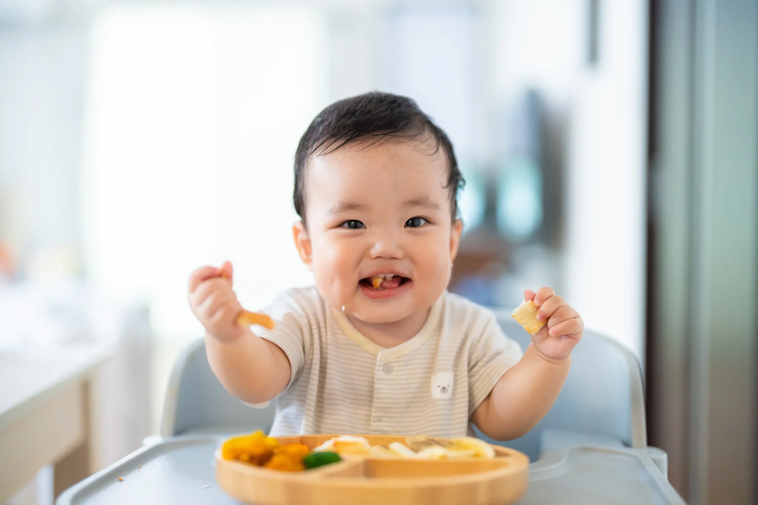 A young child with dark hair and a striped shirt eating food, smiling, sitting in a high chair with a meal tray in front of him.