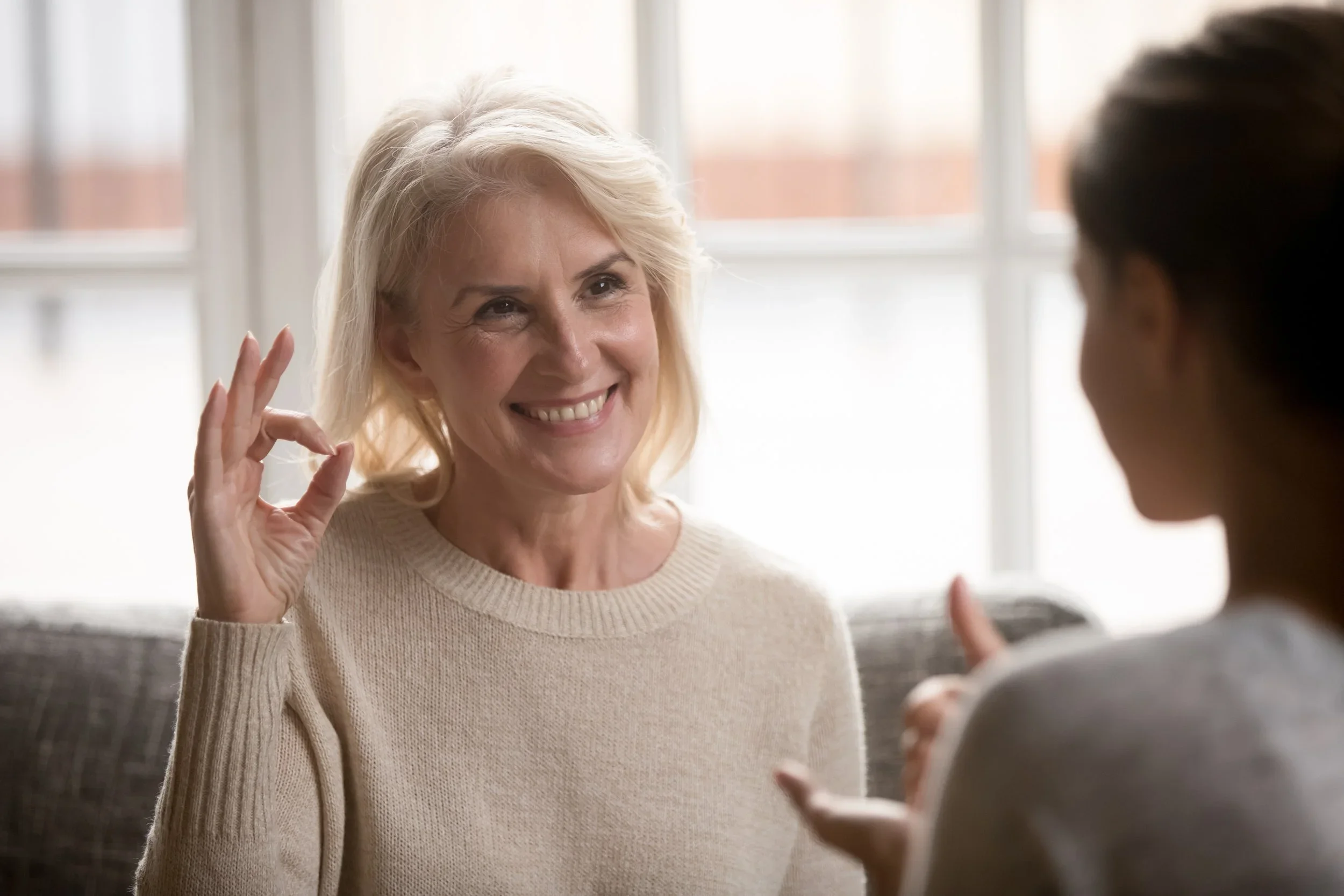 A middle-aged woman with blonde hair smiling and making an OK hand gesture, engaged in conversation with a person whose face is not visible.