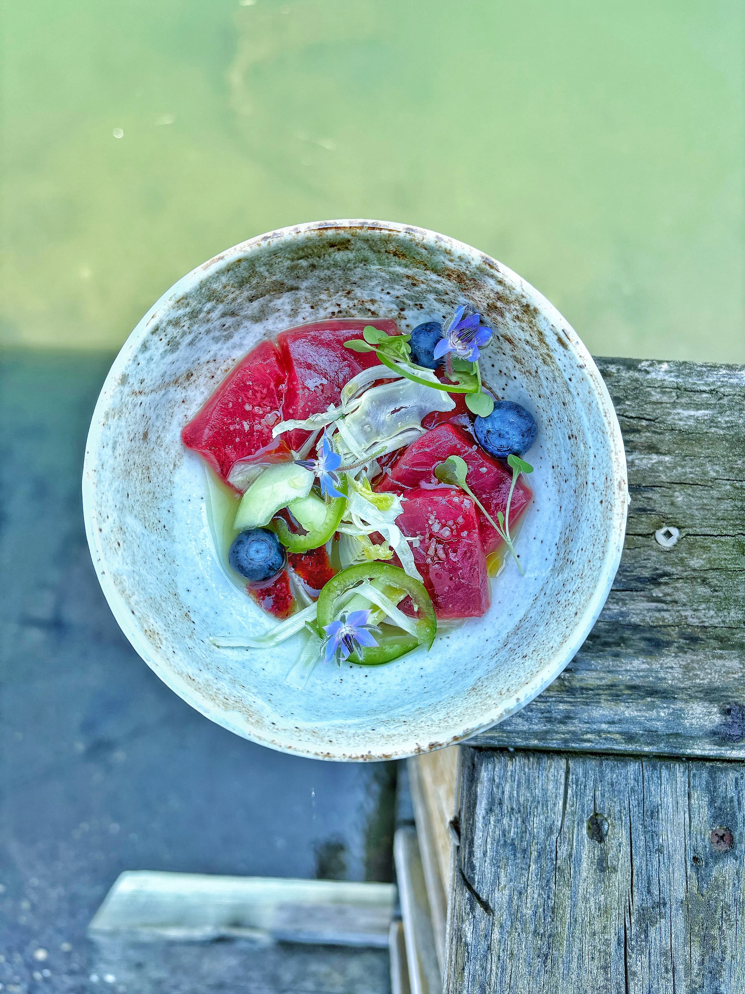 A bowl of watermelon cubes, blueberries, green cucumber slices, and edible flowers on a wooden surface outdoors.