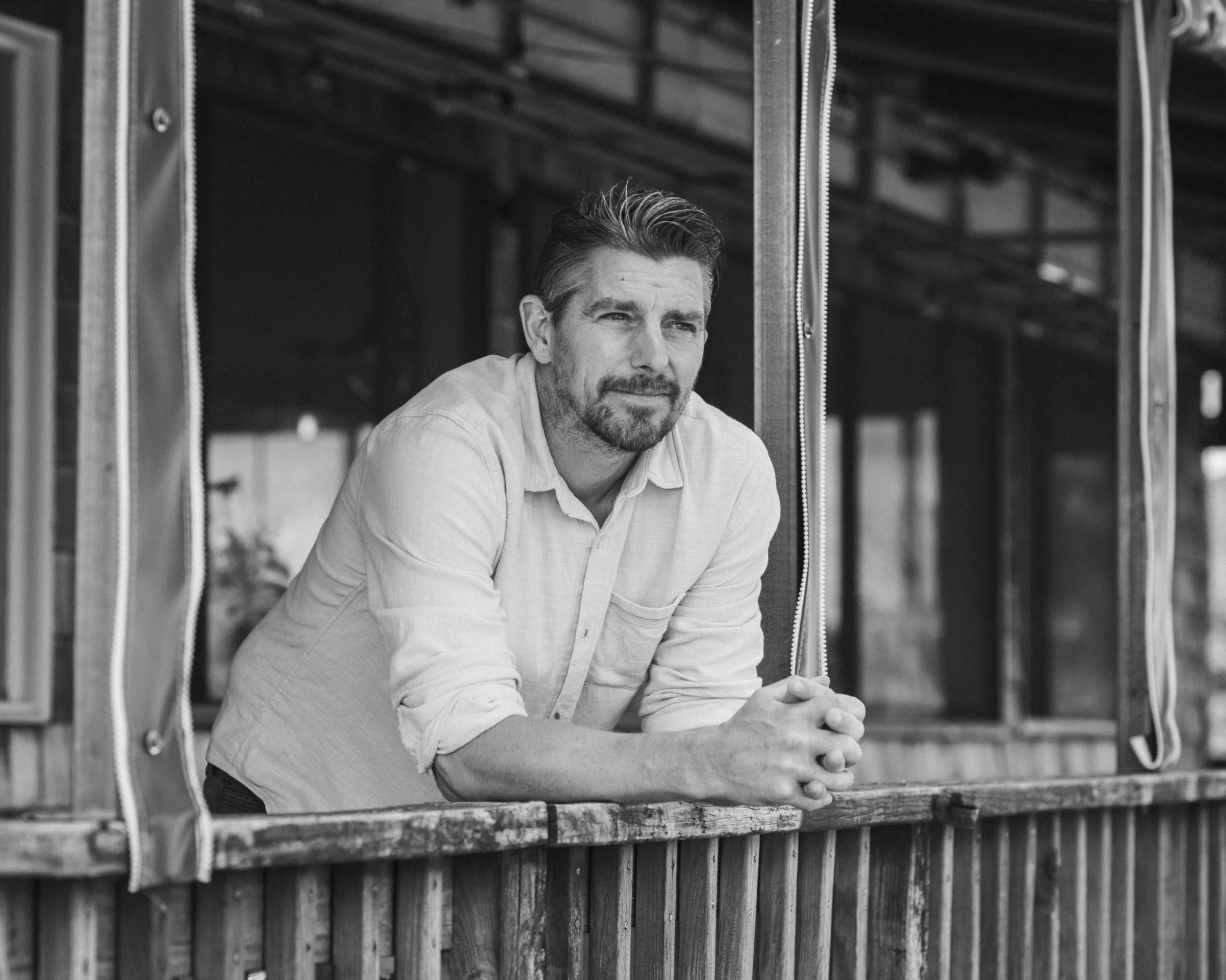 A man with a beard and mustache wearing a light-colored button-up shirt leaning on a wooden railing, looking thoughtfully into the distance.