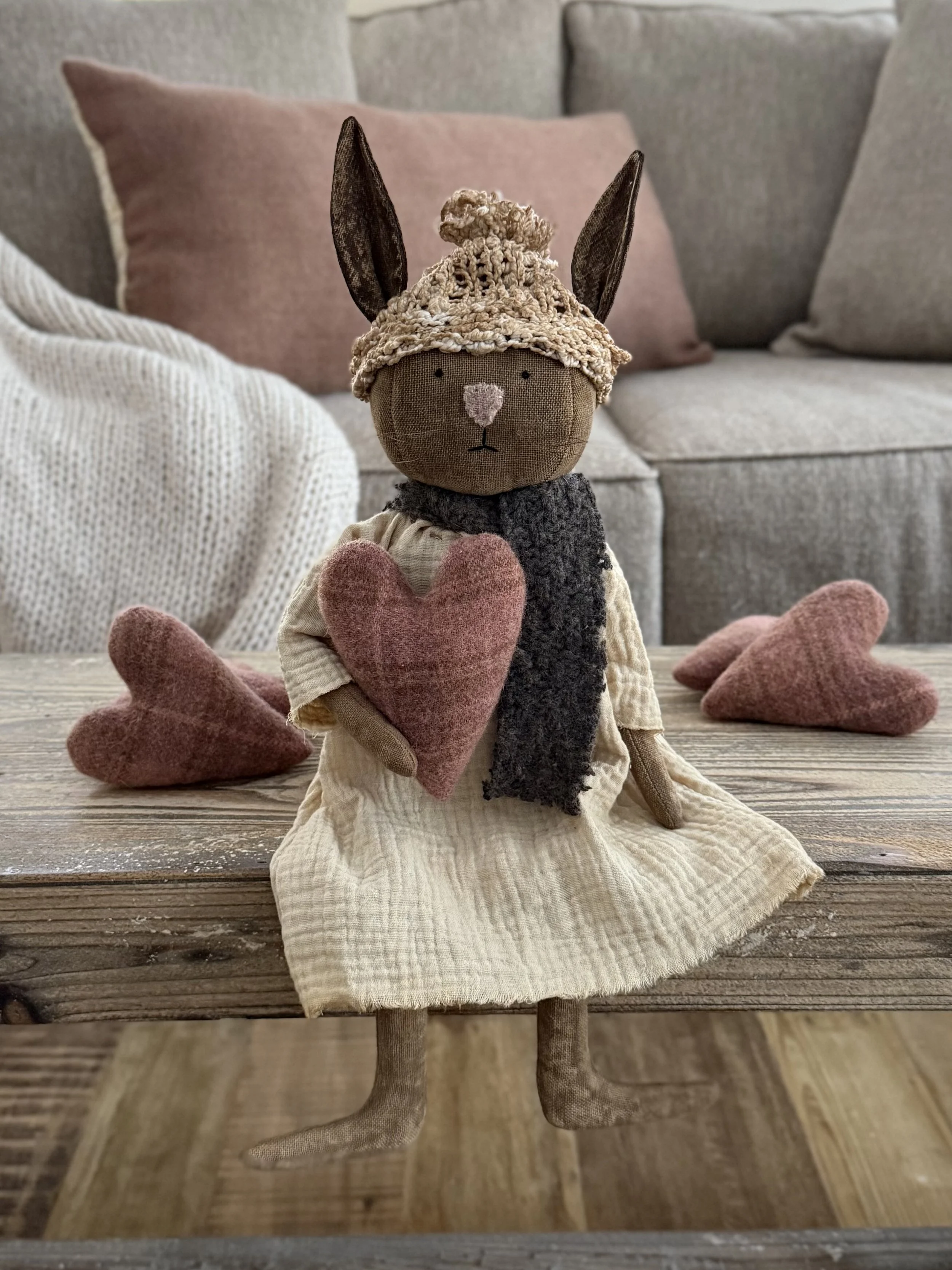 A stuffed animal bunny holding a pink heart on a wooden surface with three pink hearts around it, in a cozy living room setting.