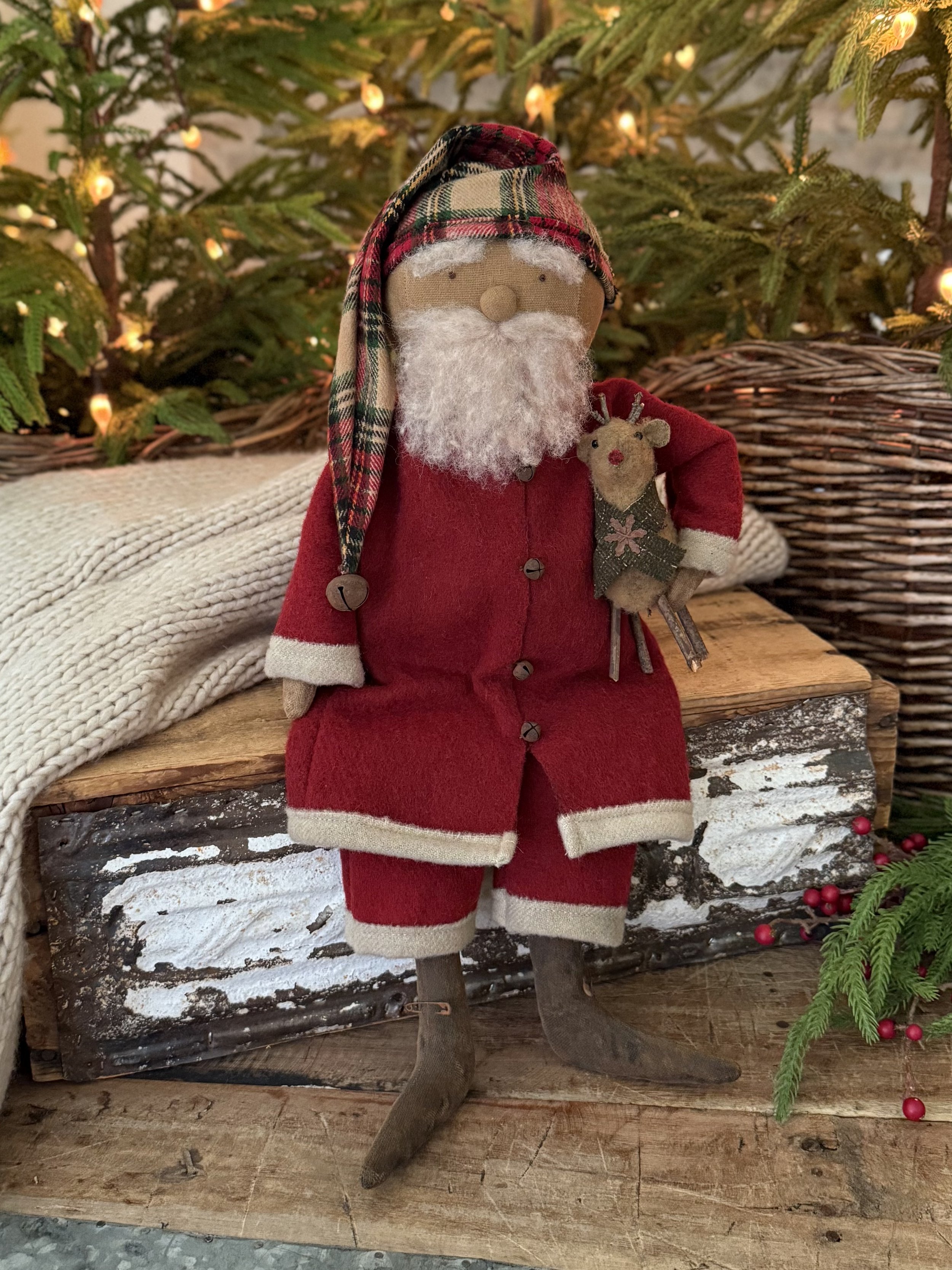 A Christmas decorative figure with a Santa Claus appearance, wearing a red coat and pants with beige trim, a plaid hat, and a fuzzy white beard. The figure has rustic, curved wooden legs and is holding a small stuffed reindeer. It is placed in front 