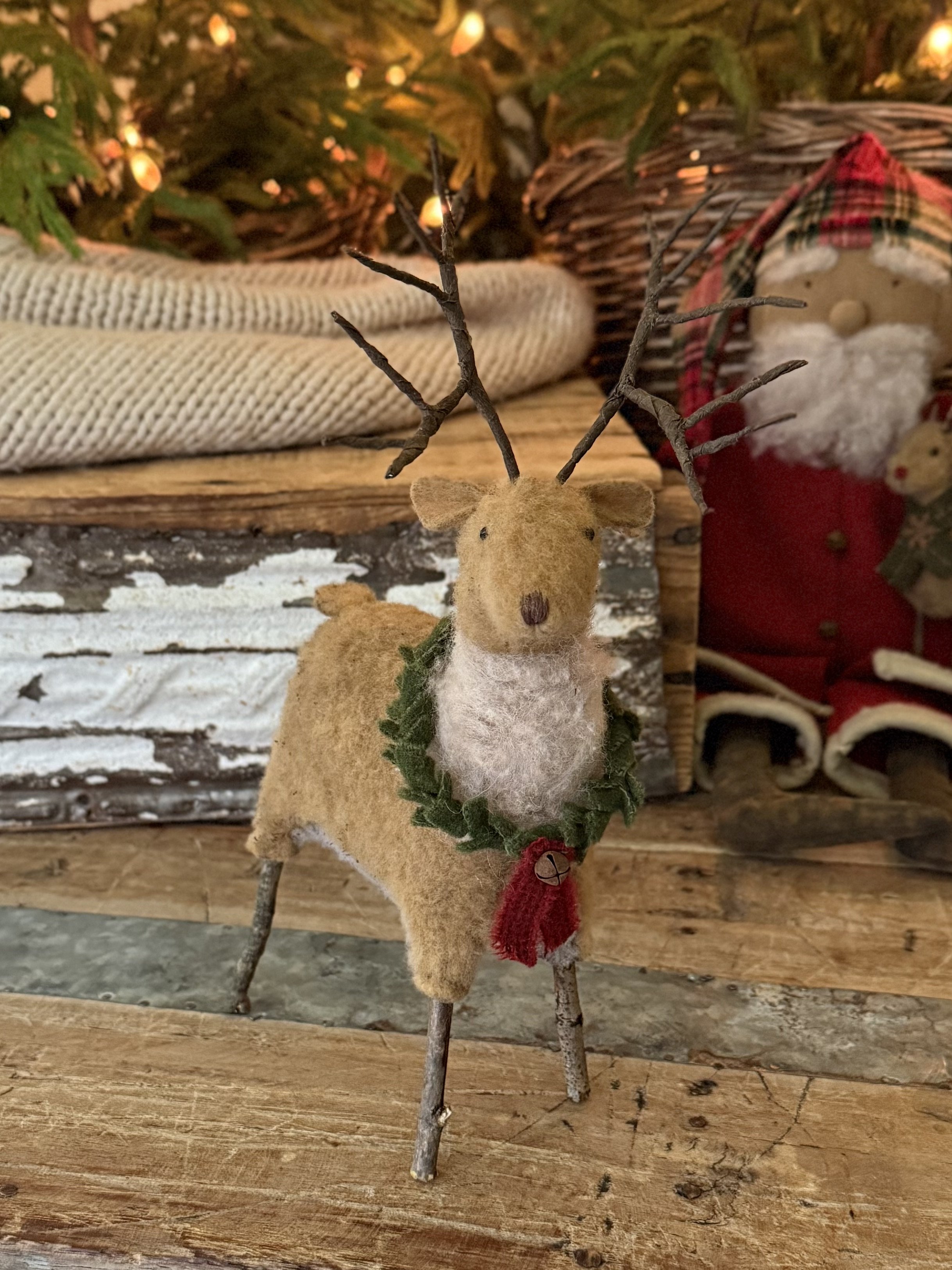 Felt reindeer decoration with twig antlers, green leafy collar with red bell, set against Christmas-themed background with Santa figure, wrapped gift, and Christmas tree lights.