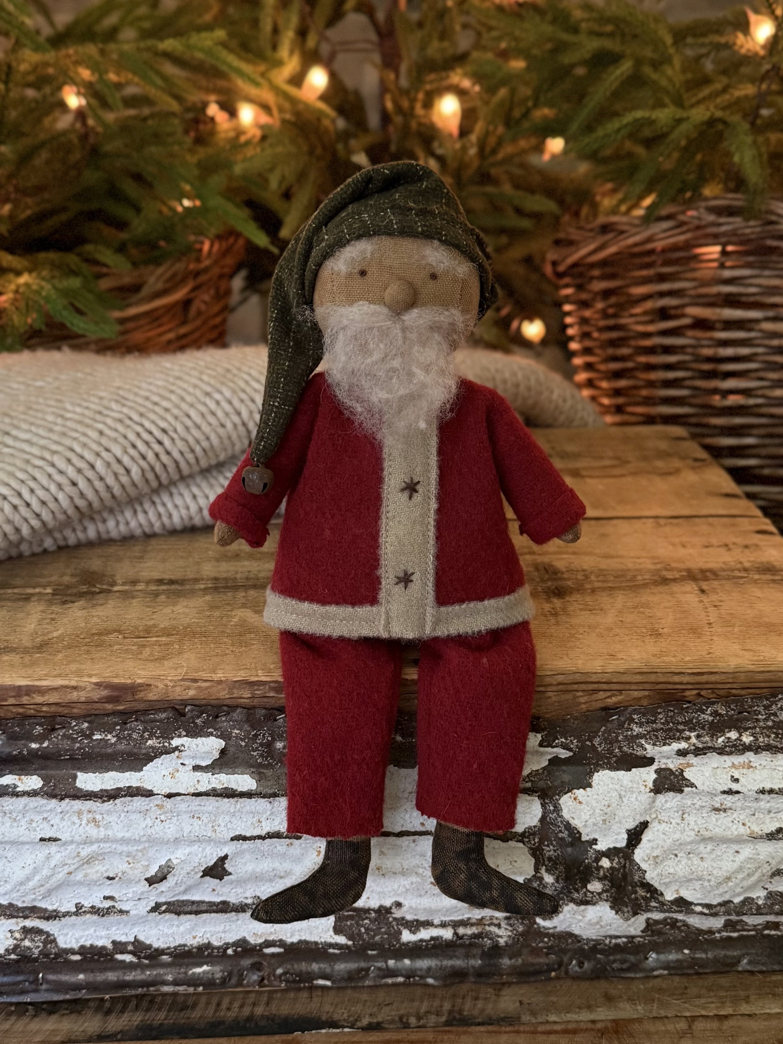A small plush Santa Claus doll with a white beard, red suit, and black boots, sitting on a wooden surface with Christmas lights and greenery in the background.