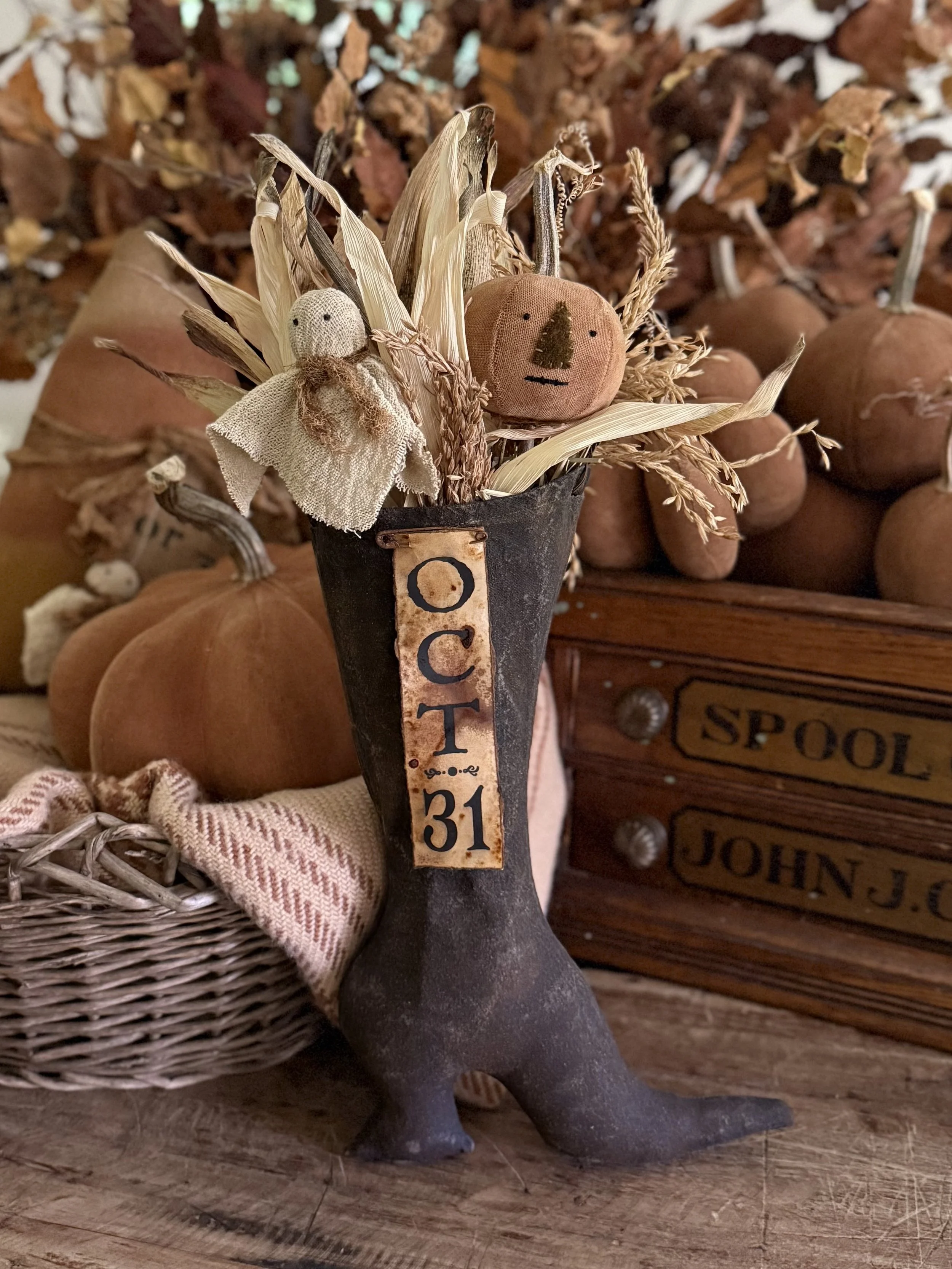 Decorative fall-themed display in a black boot-shaped container filled with dried leaves and small plush scarecrow and pumpkin ornaments. In the background, there are pumpkins, a basket with a blanket, and a wooden box labeled 'SPOOL JOHN J.'.