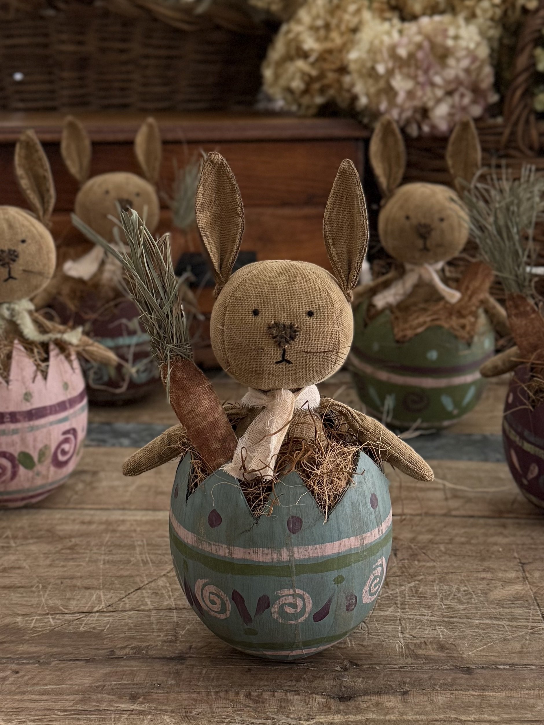 A plush bunny doll with long ears, holding a felt carrot, seated inside a decorated painted pot, with other similar bunny dolls in the background.