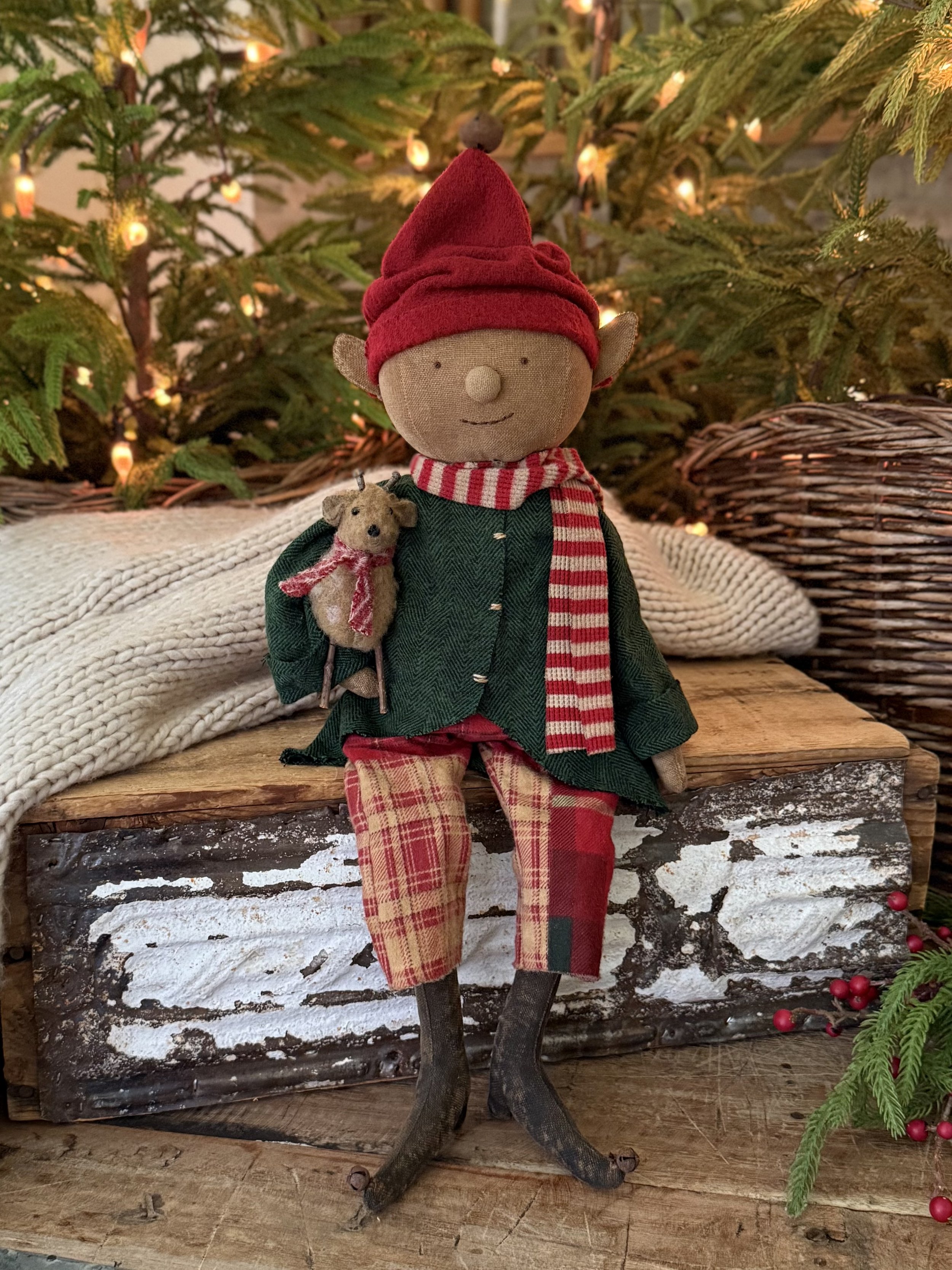 A Christmas elf doll with a round face, small ears, and a simple smile, wearing a red hat, green coat, and plaid pants, holding a small teddy bear, sitting on a rustic wooden surface in front of a decorated pine tree with lights.