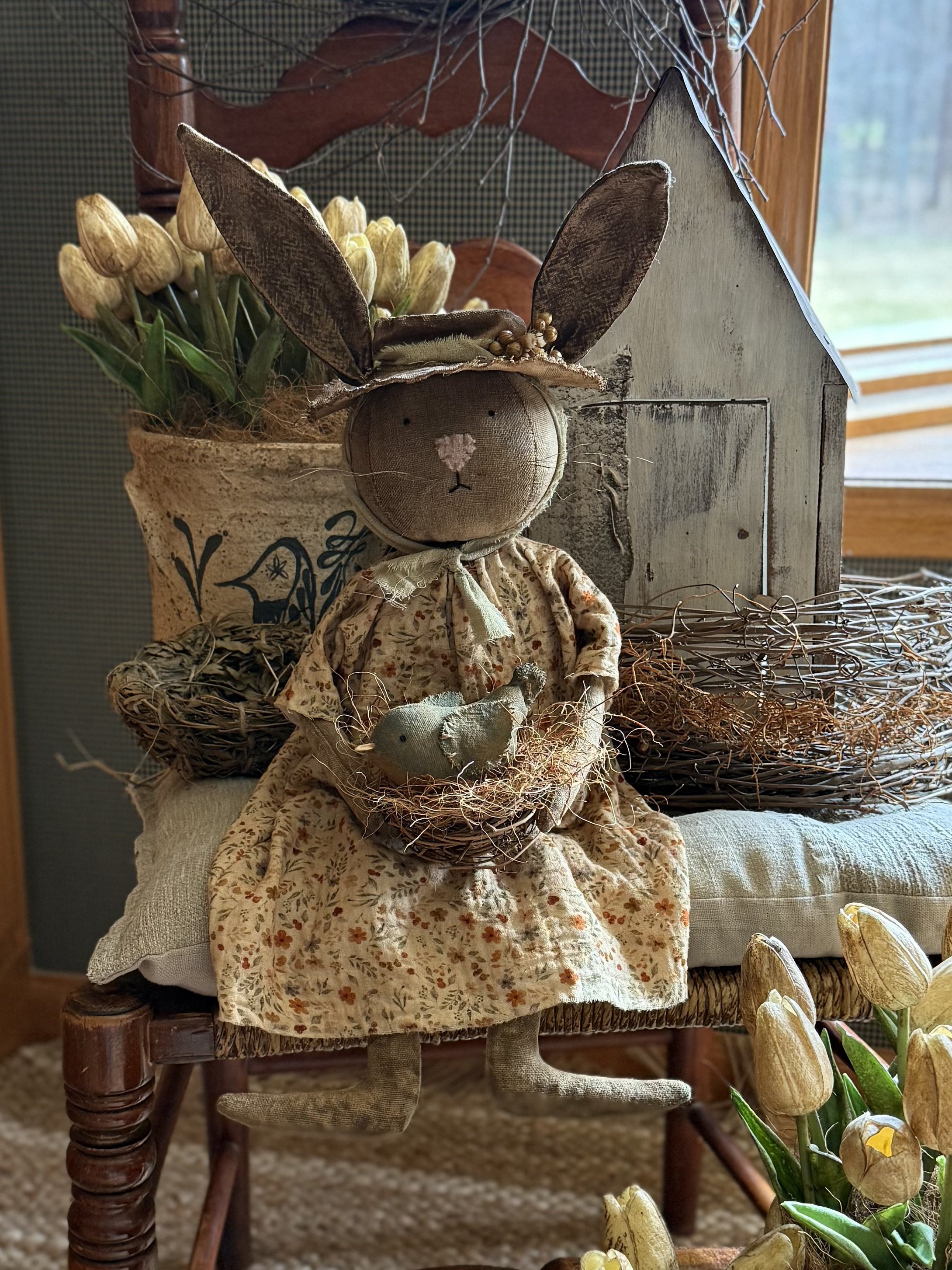 Decorative bunny doll holding a bird, sitting on a chair with floral and rustic decorations around.