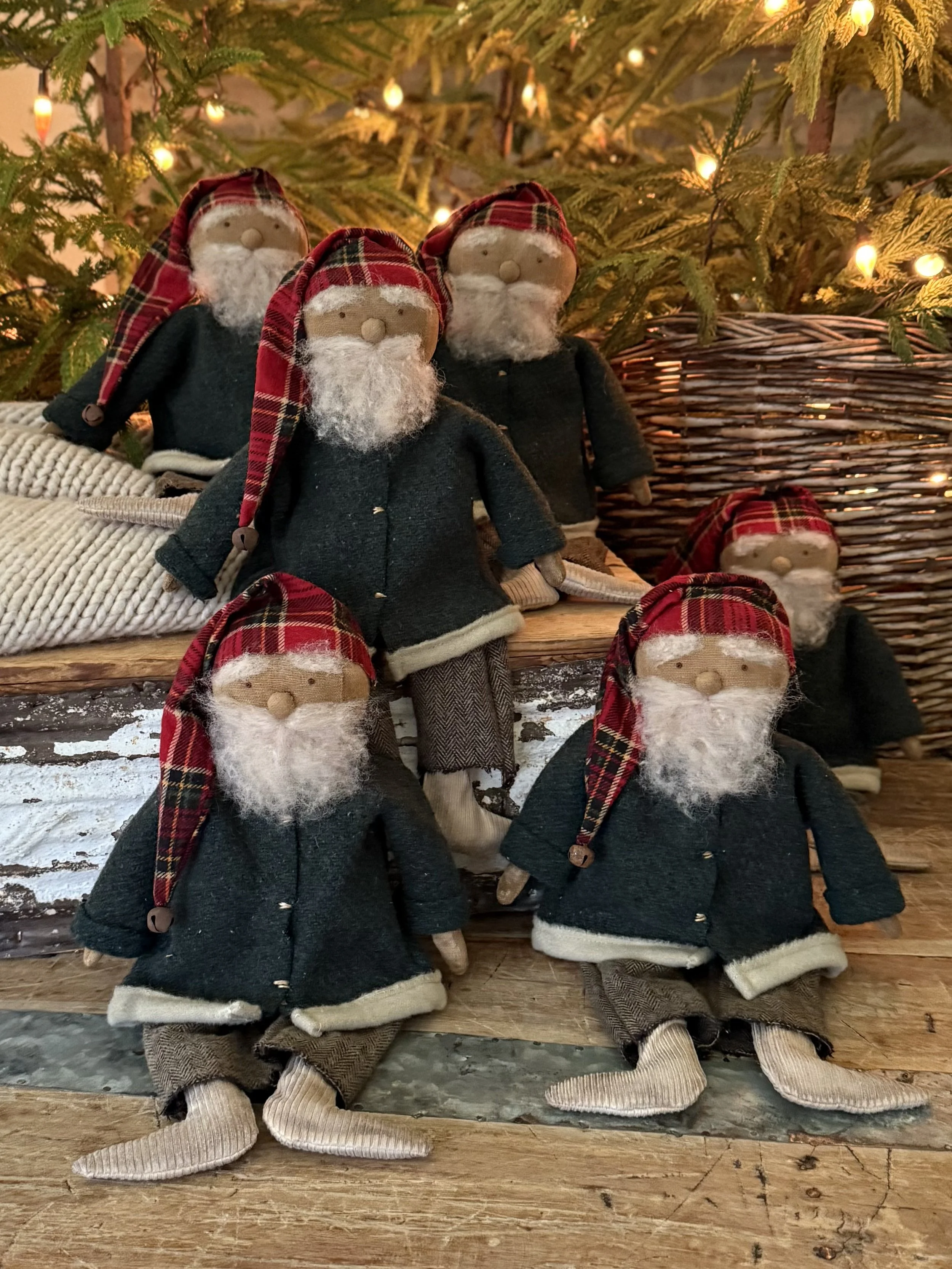 Multiple Santa Claus dolls wearing dark coats, plaid hats, and beige shoes, placed in front of a decorated Christmas tree with lights and greenery.