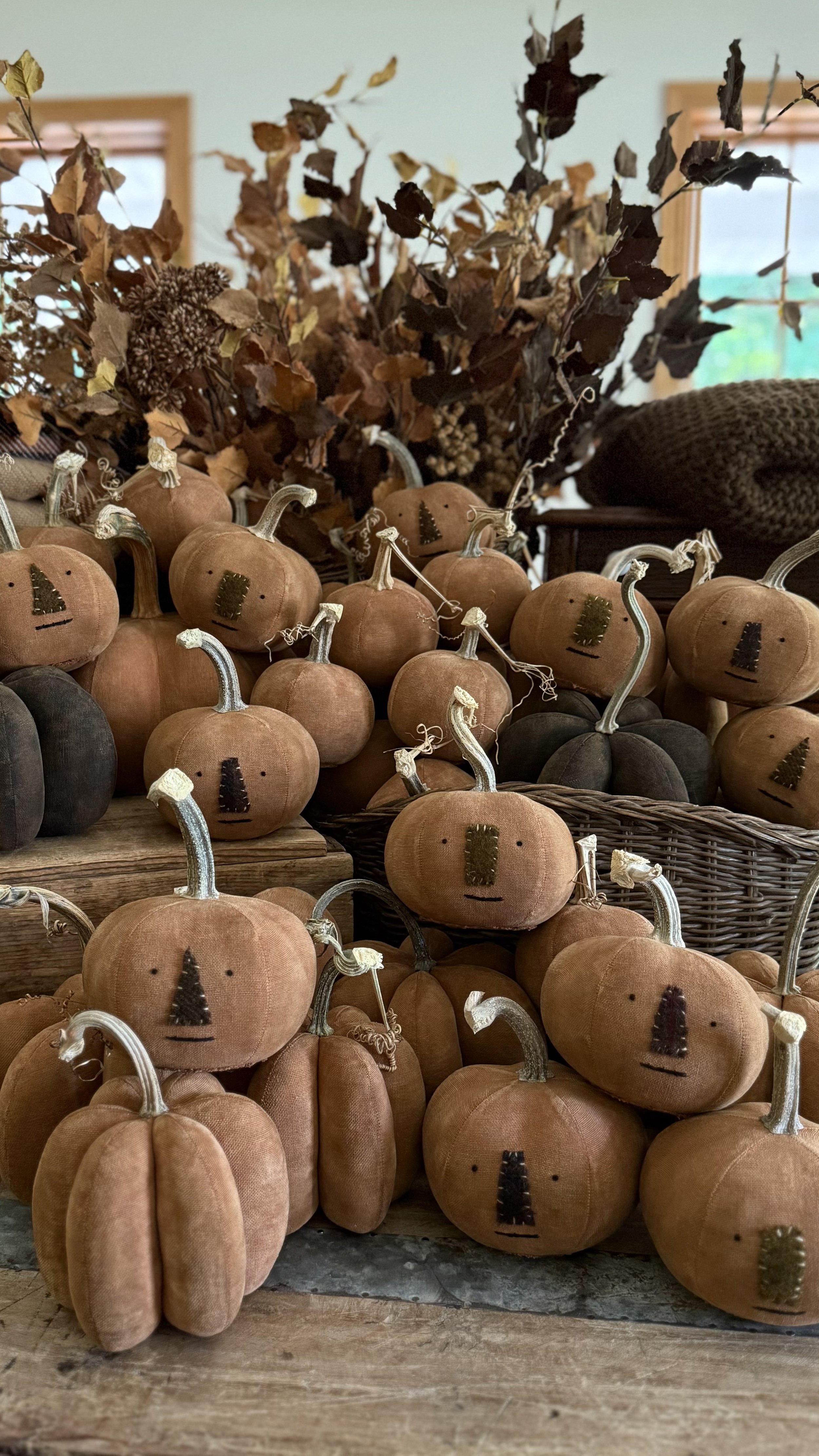 Small plush pumpkins with stitched faces and fabric leaves, surrounded by dried autumn leaves and branches, on a wooden surface.