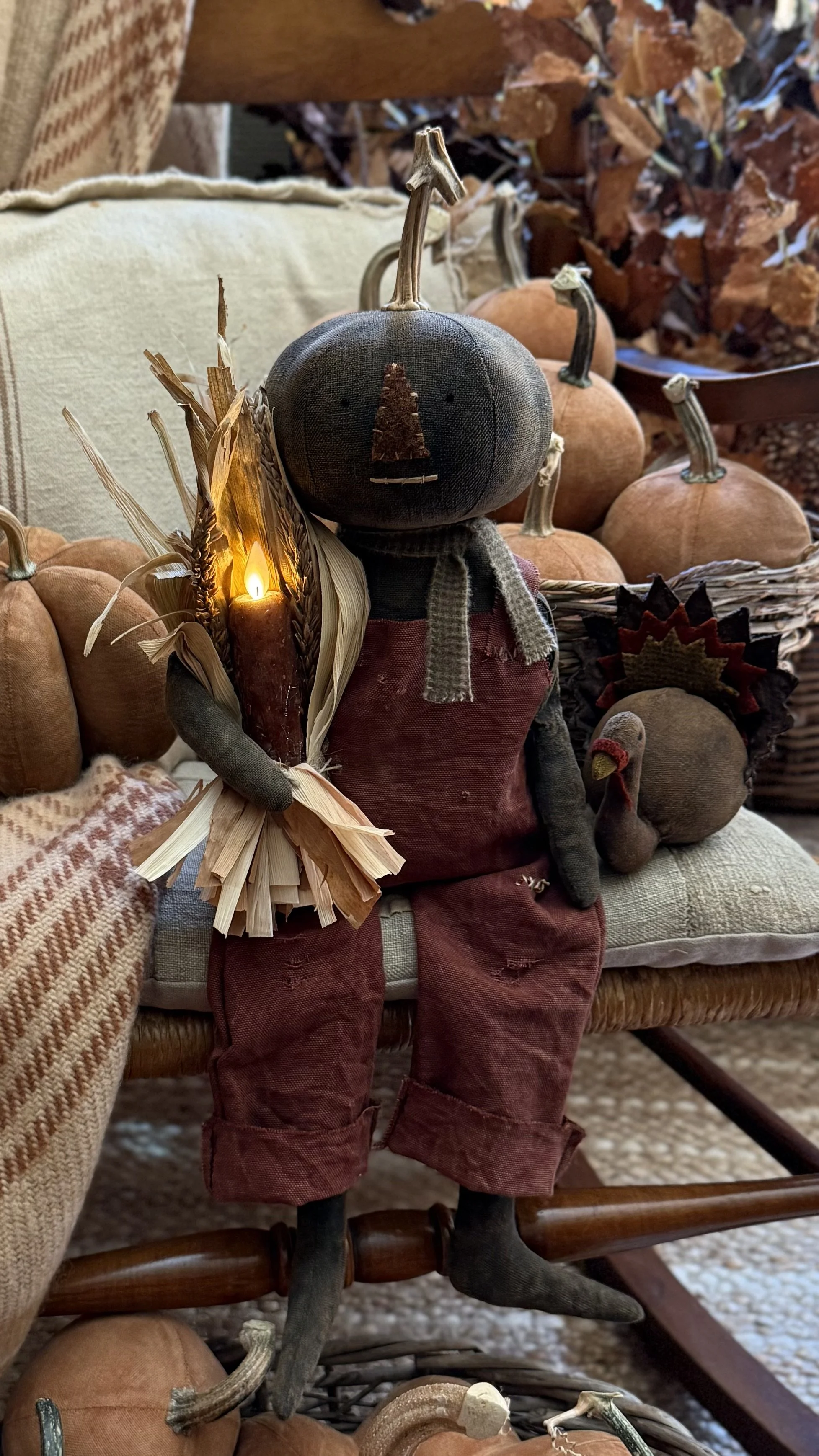 A stuffed scarecrow with a pumpkin head, wearing red overalls, holding a small illuminated torch, sitting on a bench surrounded by pumpkins and fall decorations.