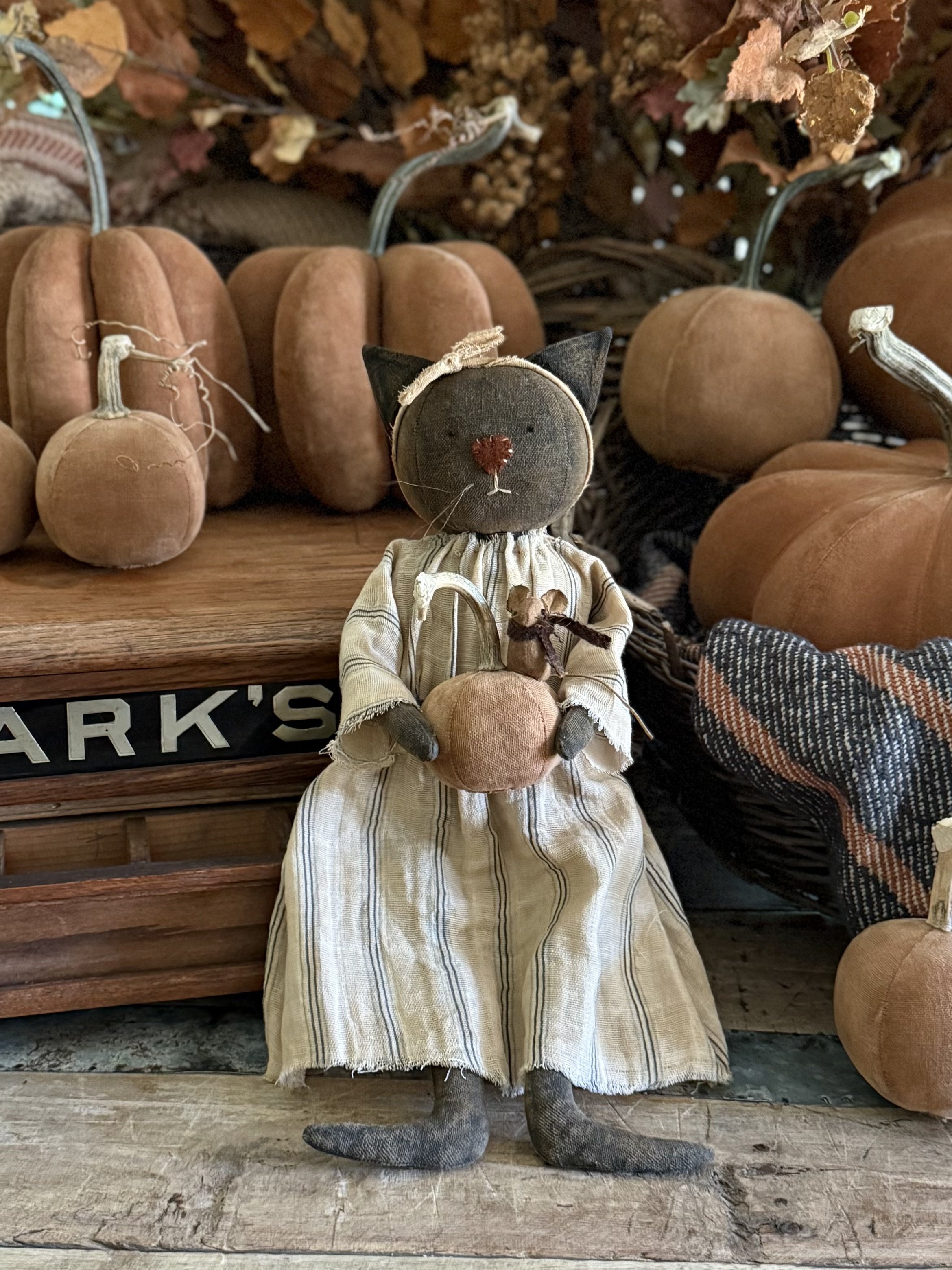 A vintage fabric cat doll dressed in a striped dress, holding a small pumpkin with a tiny mouse on its arm, surrounded by fabric pumpkins.