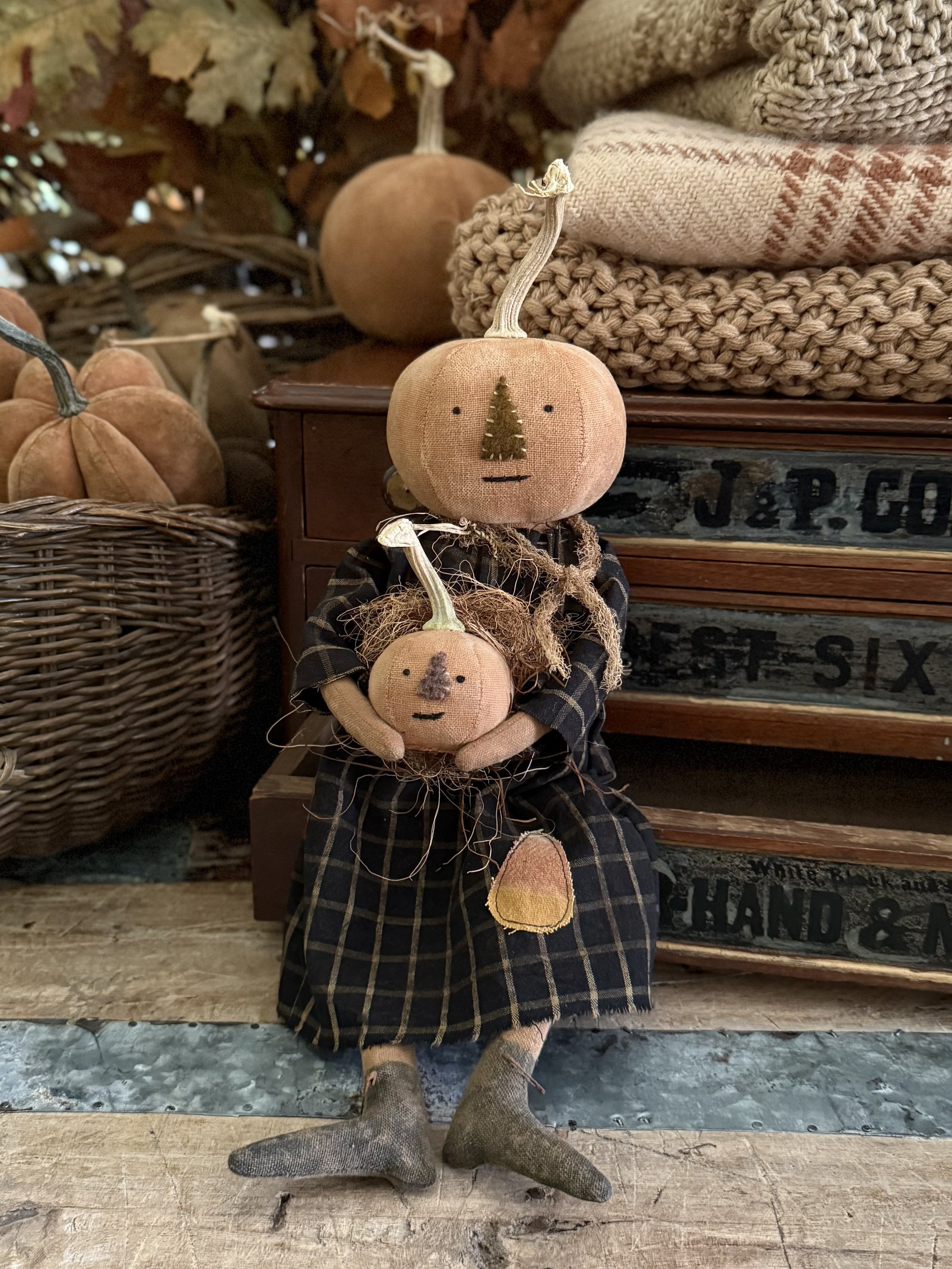 Decorative fabric scarecrows with pumpkin-shaped heads and faces, dressed in plaid and vintage clothing, set in a rustic autumn display with pumpkins and woven baskets.