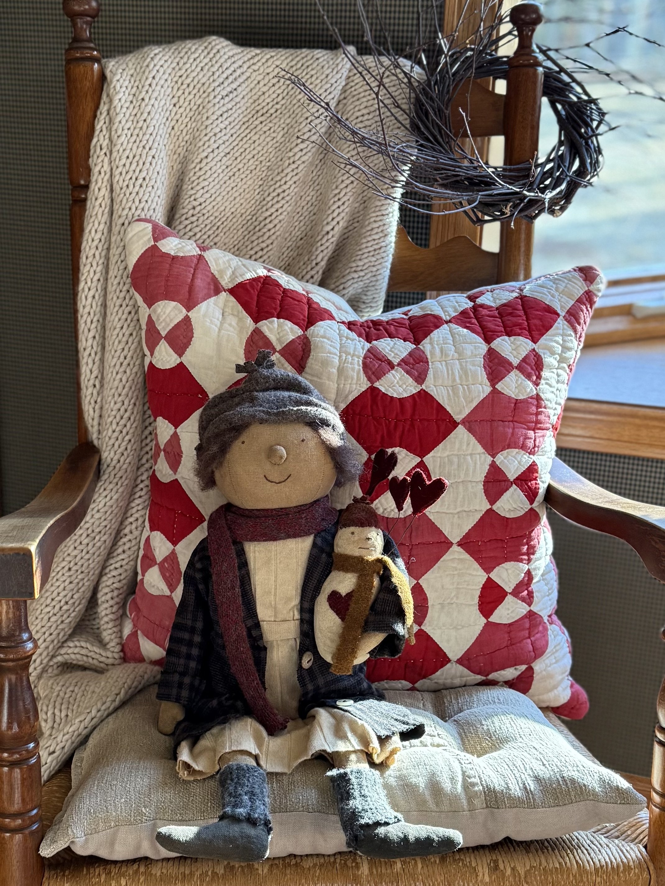 A handmade cloth doll with gray hair, wearing a gray hat, black and white checkered jacket, beige skirt, and gray socks, sitting on a cushion in front of a red and white quilted pillow. The doll is holding a smaller doll with a brown scarf, and there