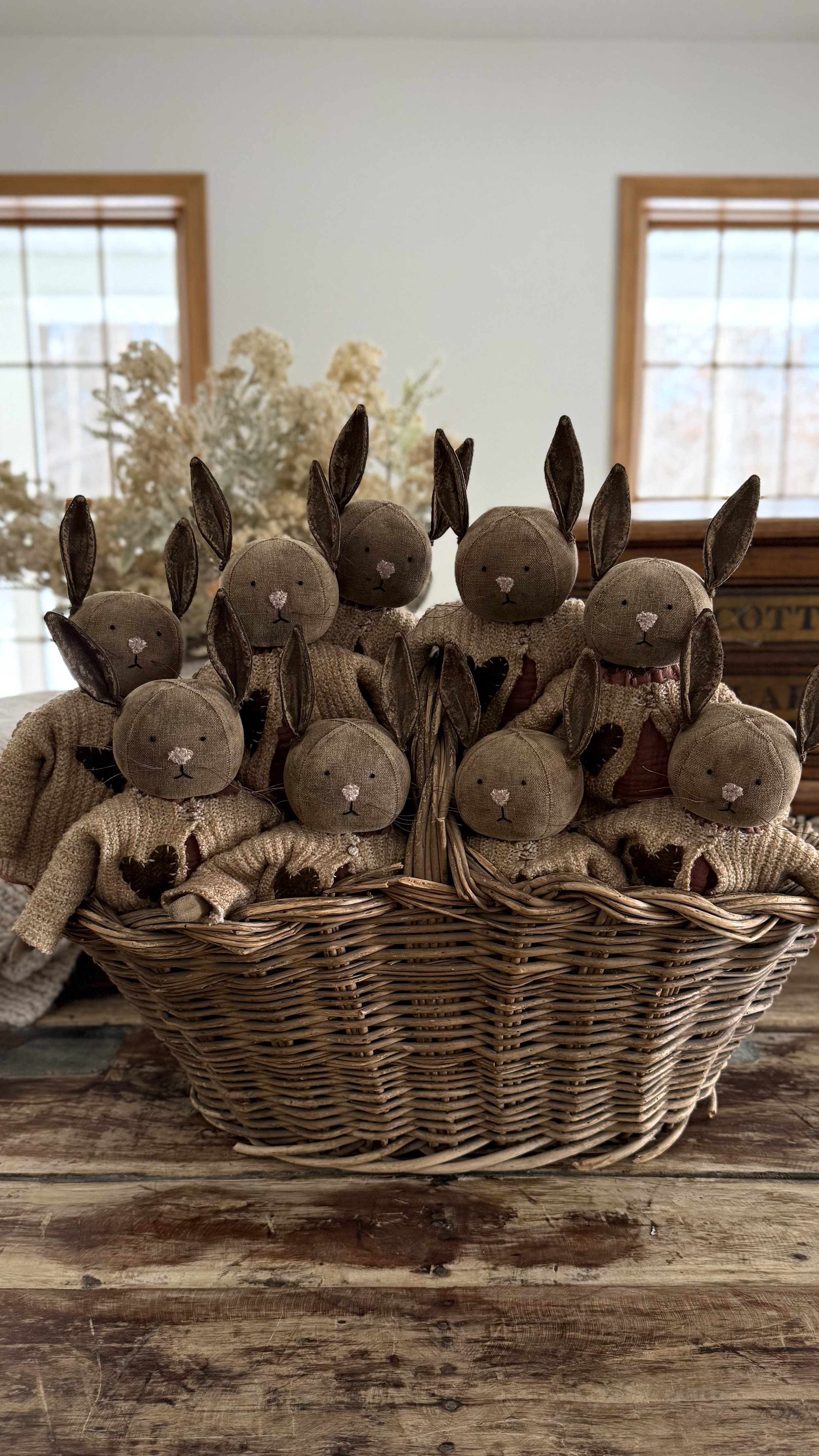 A basket filled with rustic stuffed bunny toys with fabric ears, noses, and limbs, sitting on a wooden table, with a background of windows and dried flowers.