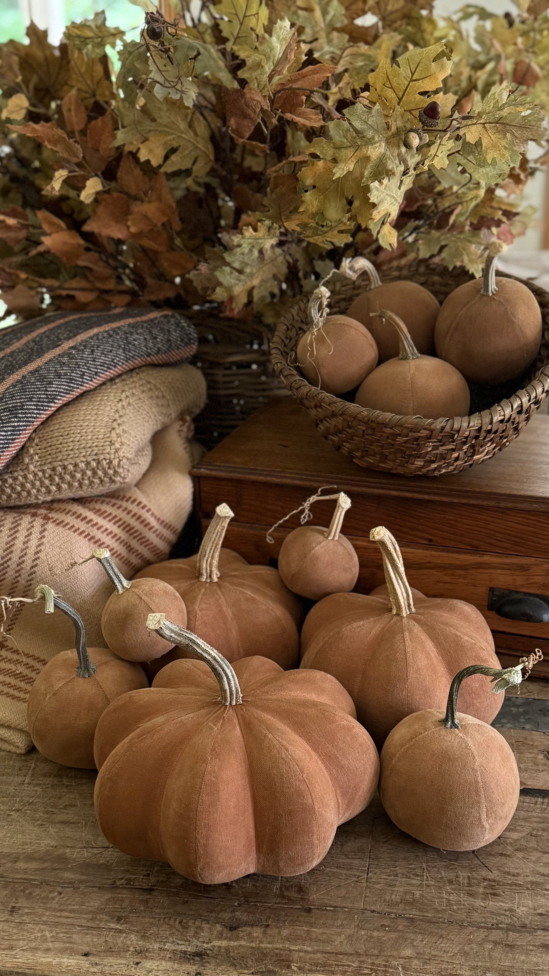 Arrangement of fabric pumpkins on a wooden surface with fall leaves and textiles in the background.