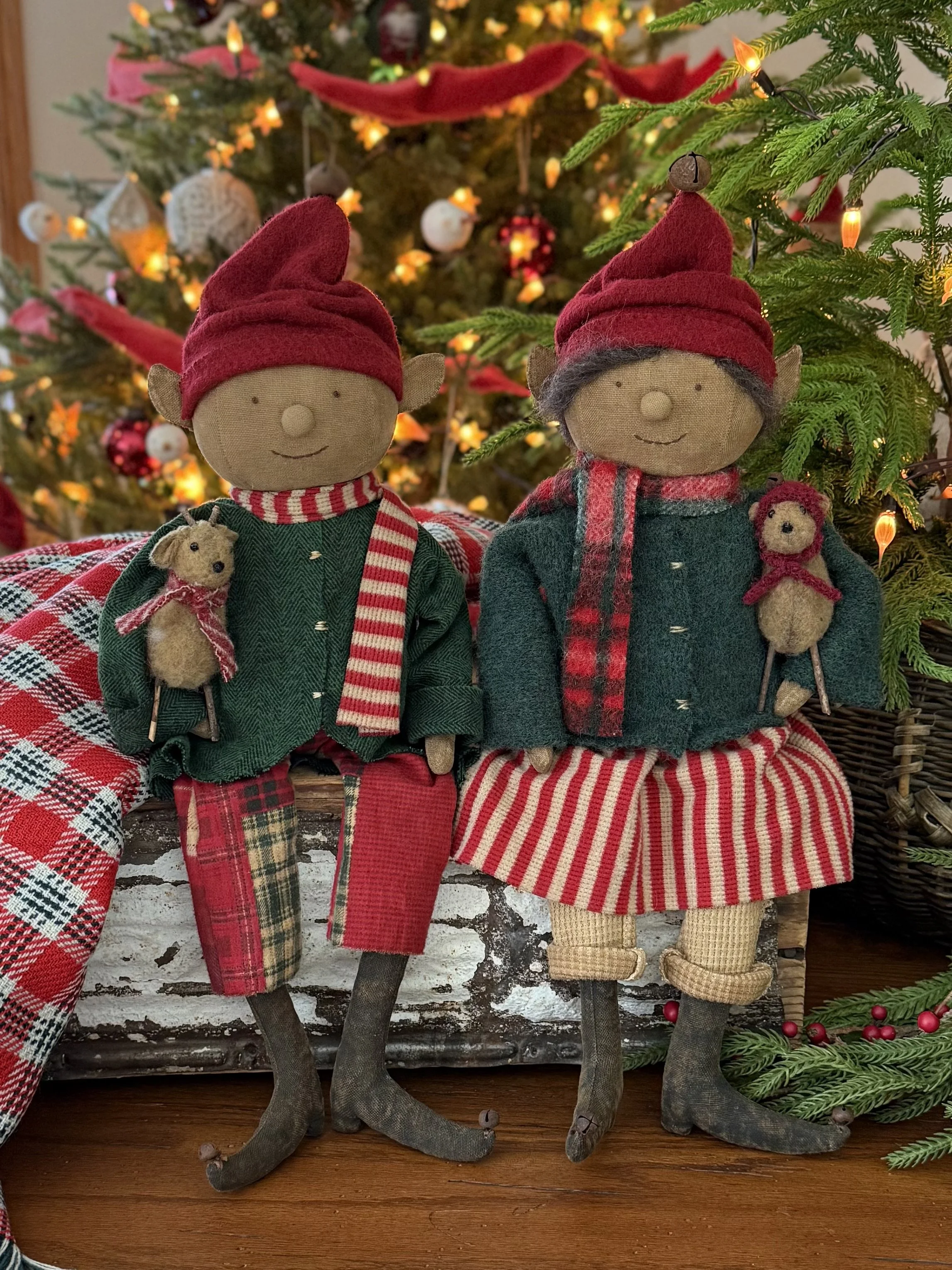 Two Christmas elf dolls with red hats, green jackets, and plaid pants holding small teddy bear toys, standing in front of a decorated Christmas tree with lights and ornaments.