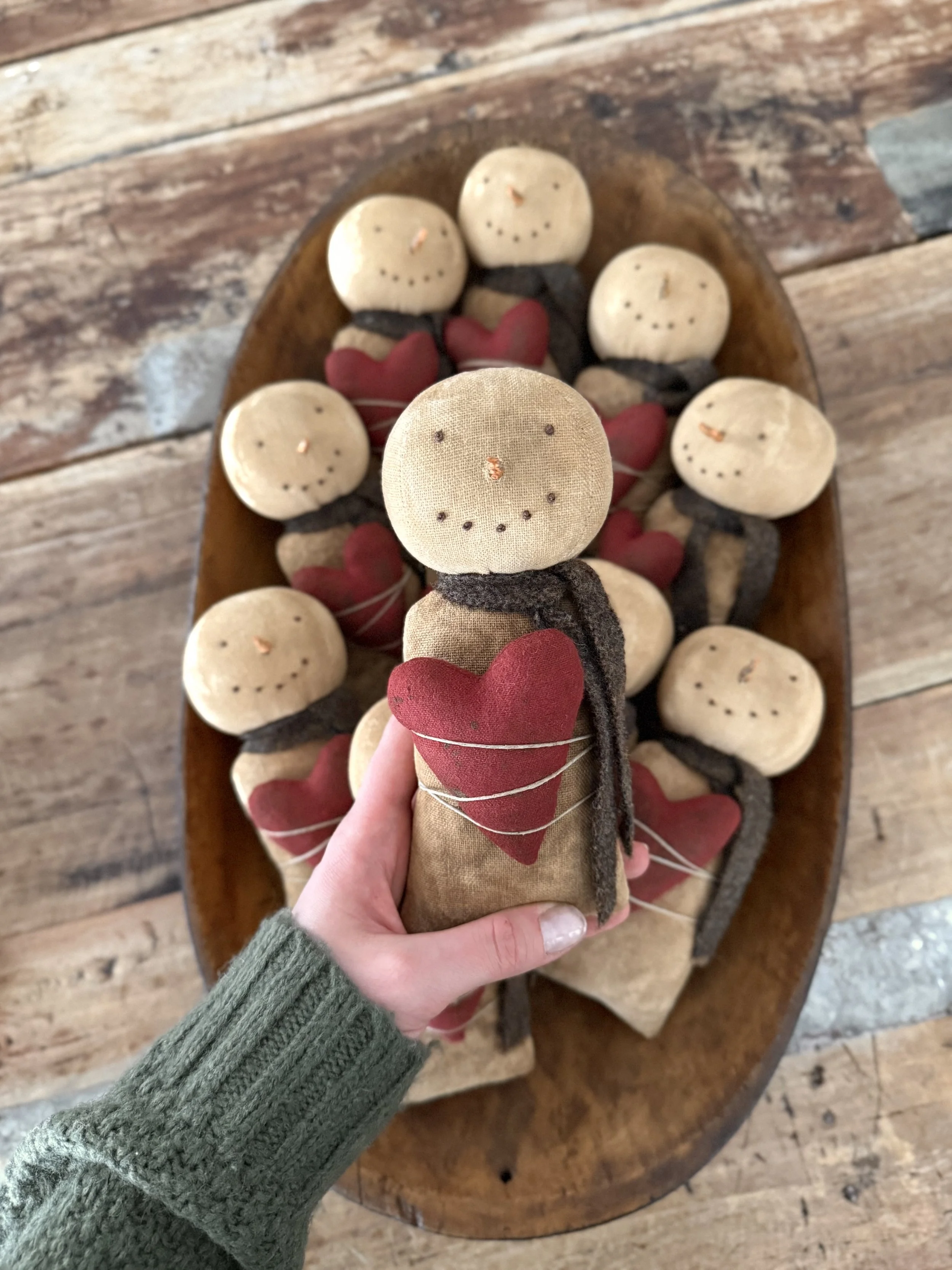 A person holding a handmade fabric doll with a smiling face made of dots, a red heart on its chest, and a dark scarf around its neck. In the background, there are several similar dolls lying on a wooden oval tray.