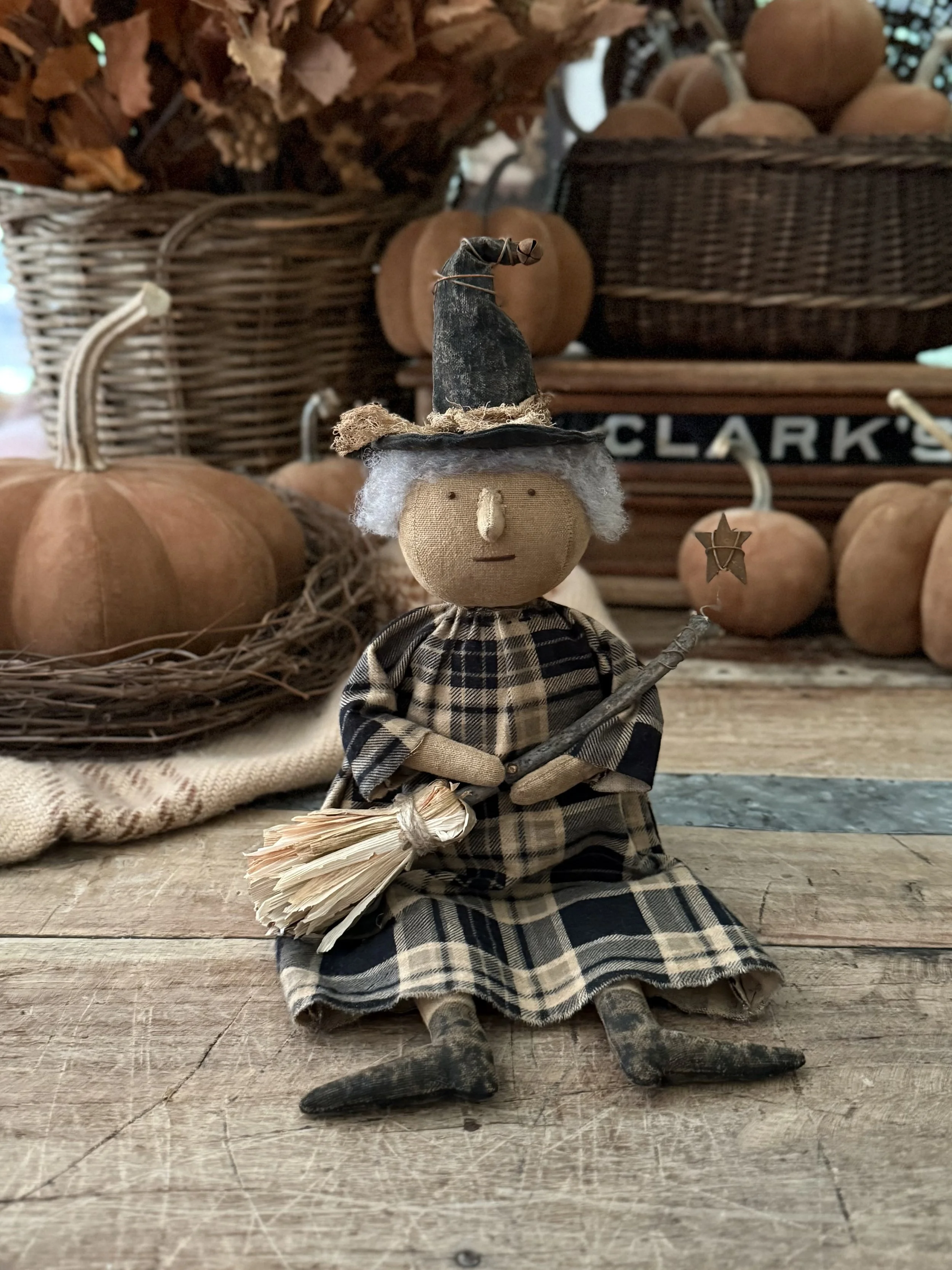 Decorative witch doll with a pointed hat holding a tiny star on a stick, sitting on a wooden surface surrounded by pumpkins and autumn-themed decor.