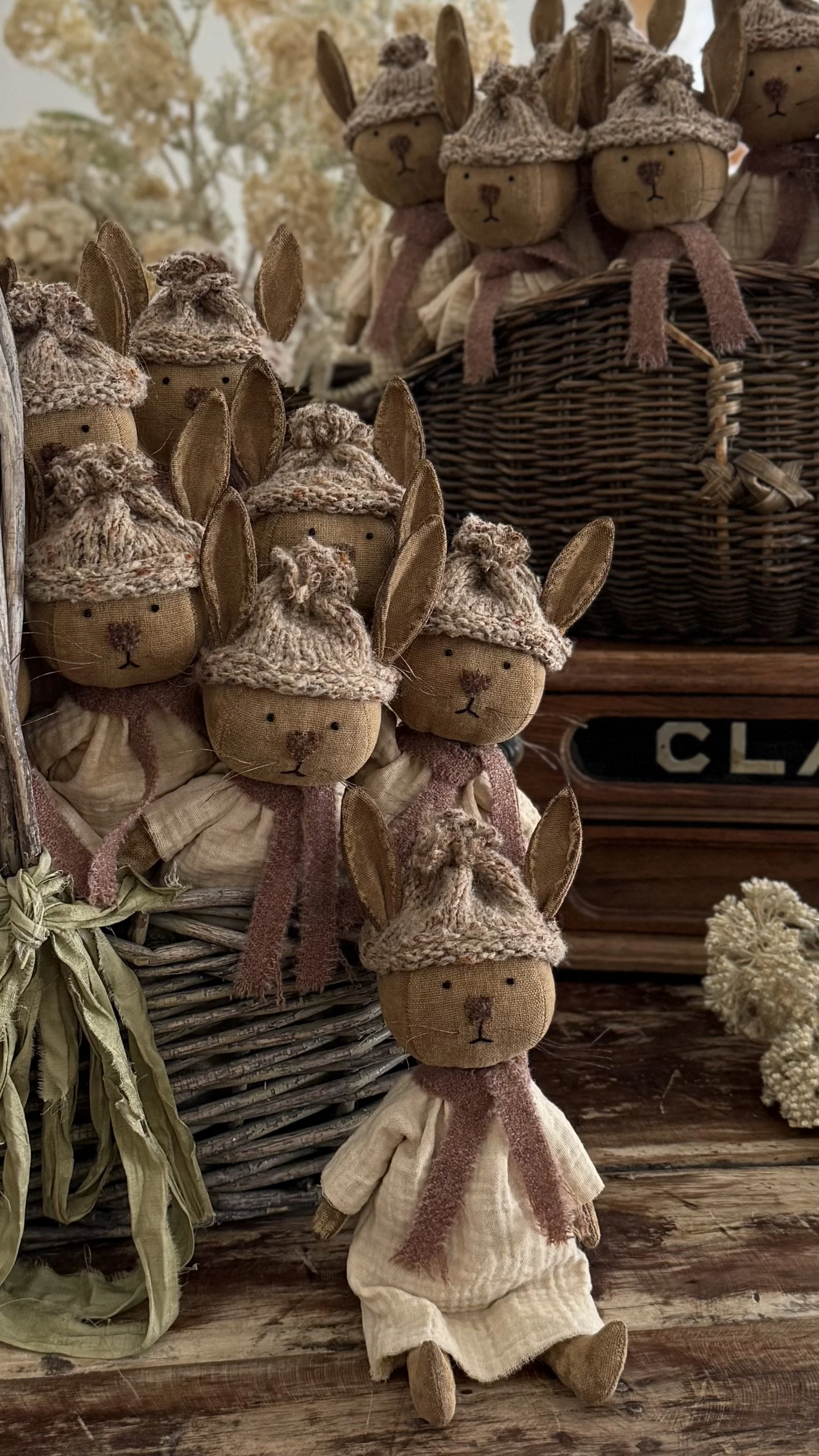 A collection of plush bunny dolls with fabric ears, wearing cozy outfits and knit hats, arranged in a wicker basket and standing on a rustic wooden surface.