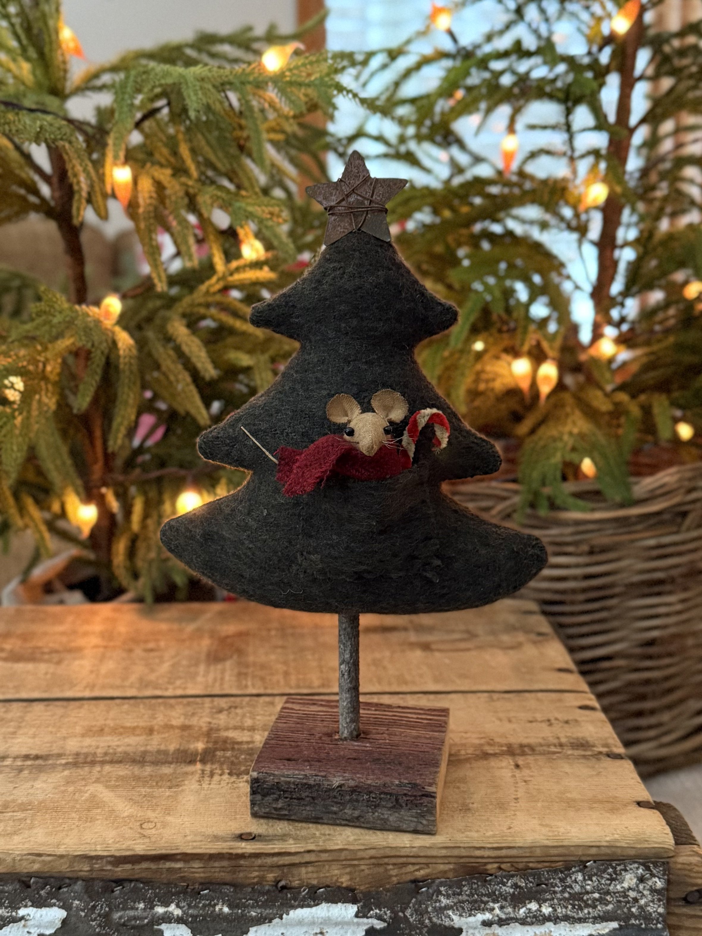 Decorative Christmas tree ornament featuring a felt Christmas tree with a mouse holding a candy cane and wearing a red scarf, standing on a wooden base in front of a lit Christmas tree with string lights.
