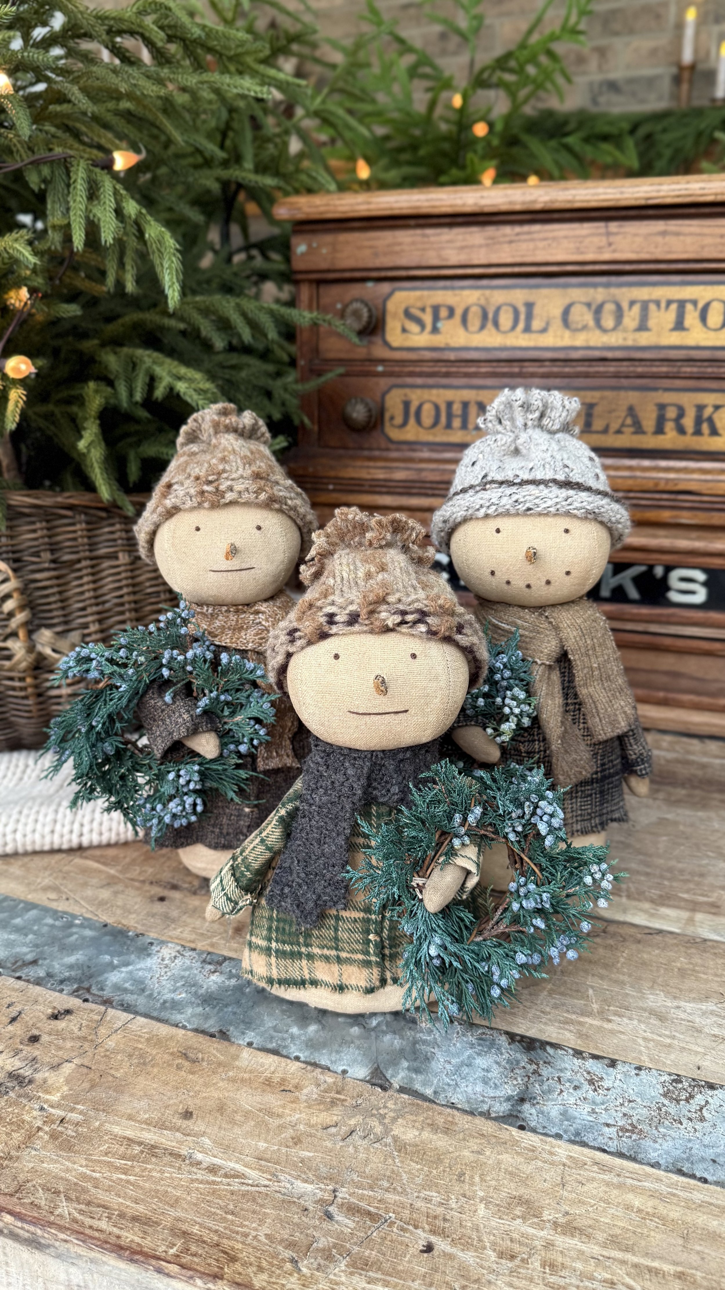 Three fabric dolls dressed in winter clothing, each holding a small wreath, standing in front of a Christmas tree and a wooden box with vintage labeling.
