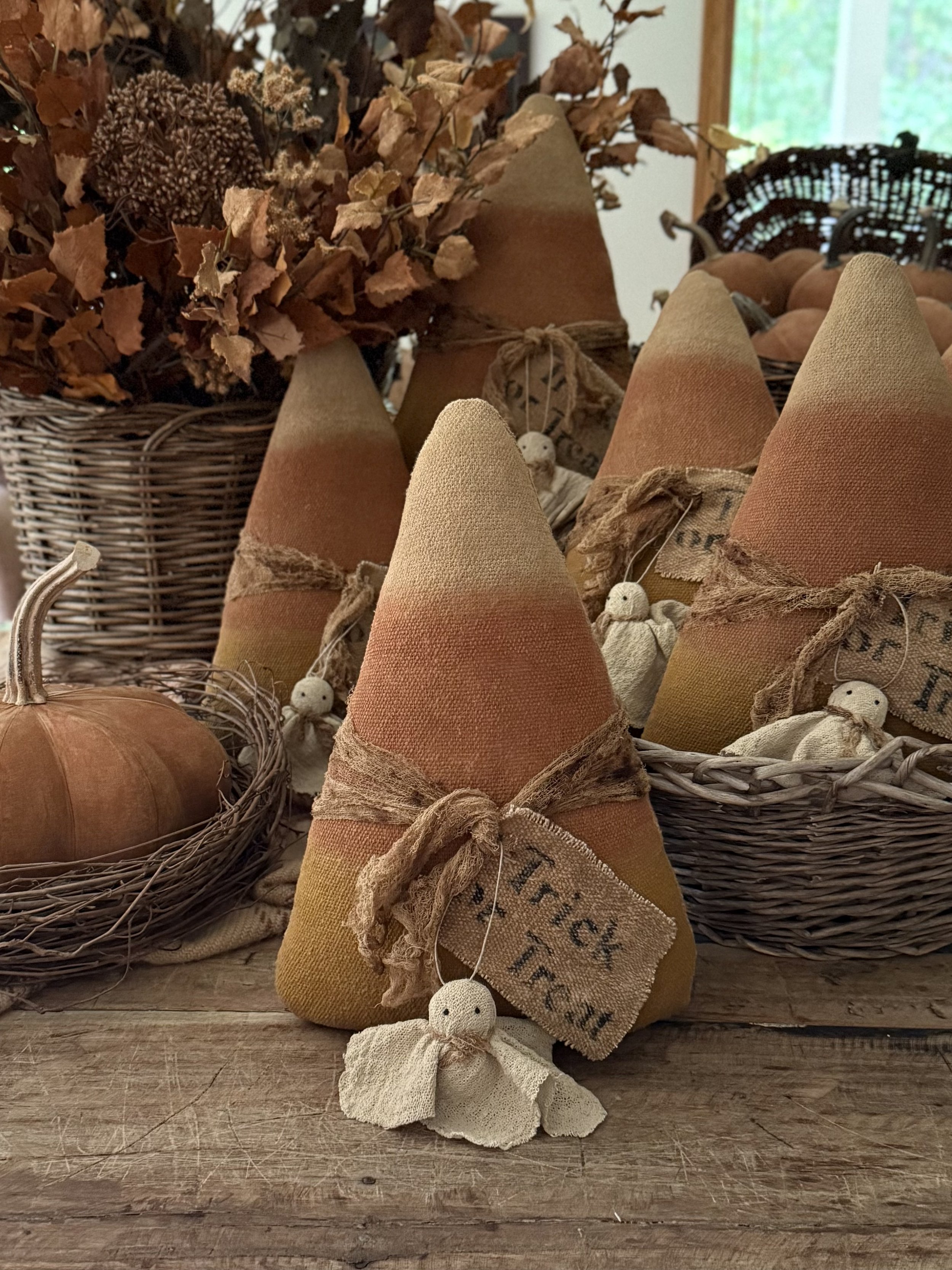 Fall-themed display with fabric cone decorations, small stuffed dolls, a pumpkin, dried flowers, and basket of pumpkins on a wooden surface.