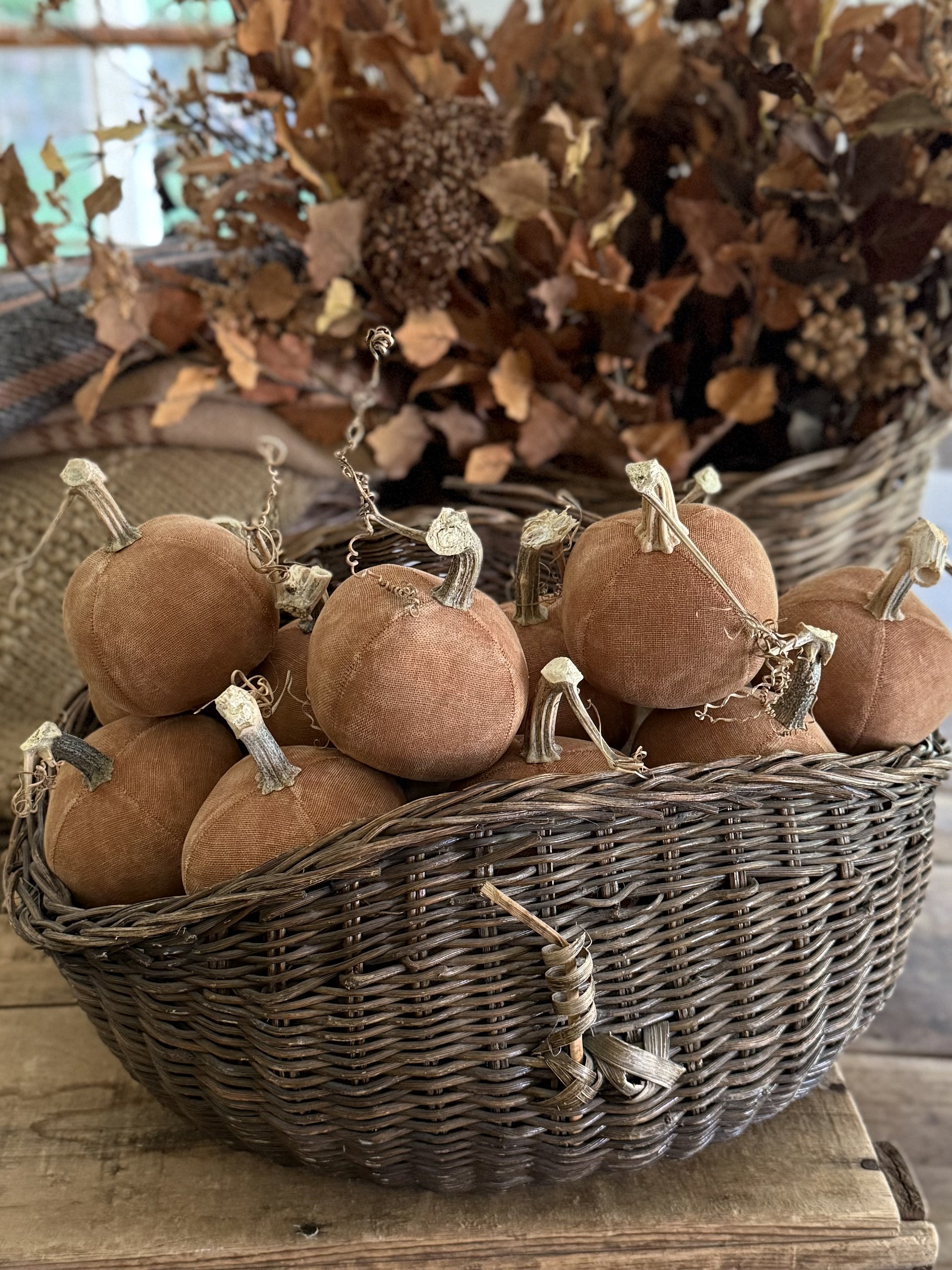 A wicker basket filled with small, fabric-covered pumpkins in earthy tones with dried stems, placed on a wooden surface with a background of a large basket of dried leaves and flowers.