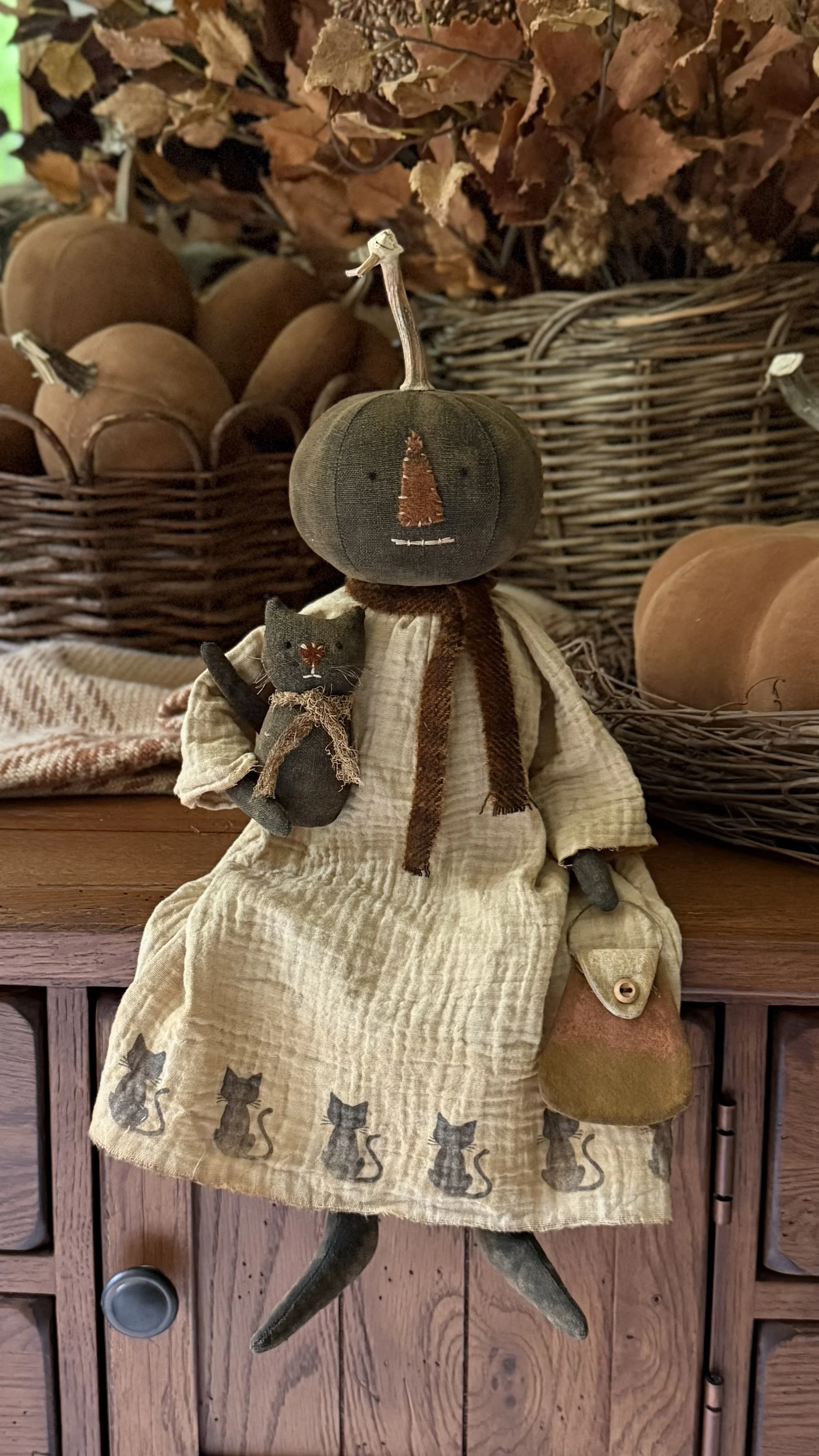 A fabric scarecrow decoration with a pumpkin head, orange stitched nose, and a brown scarf. It is holding a small black cat plush and wearing a beige dress with black cat silhouettes at the hem. The scene includes baskets of pumpkins and dried leaves