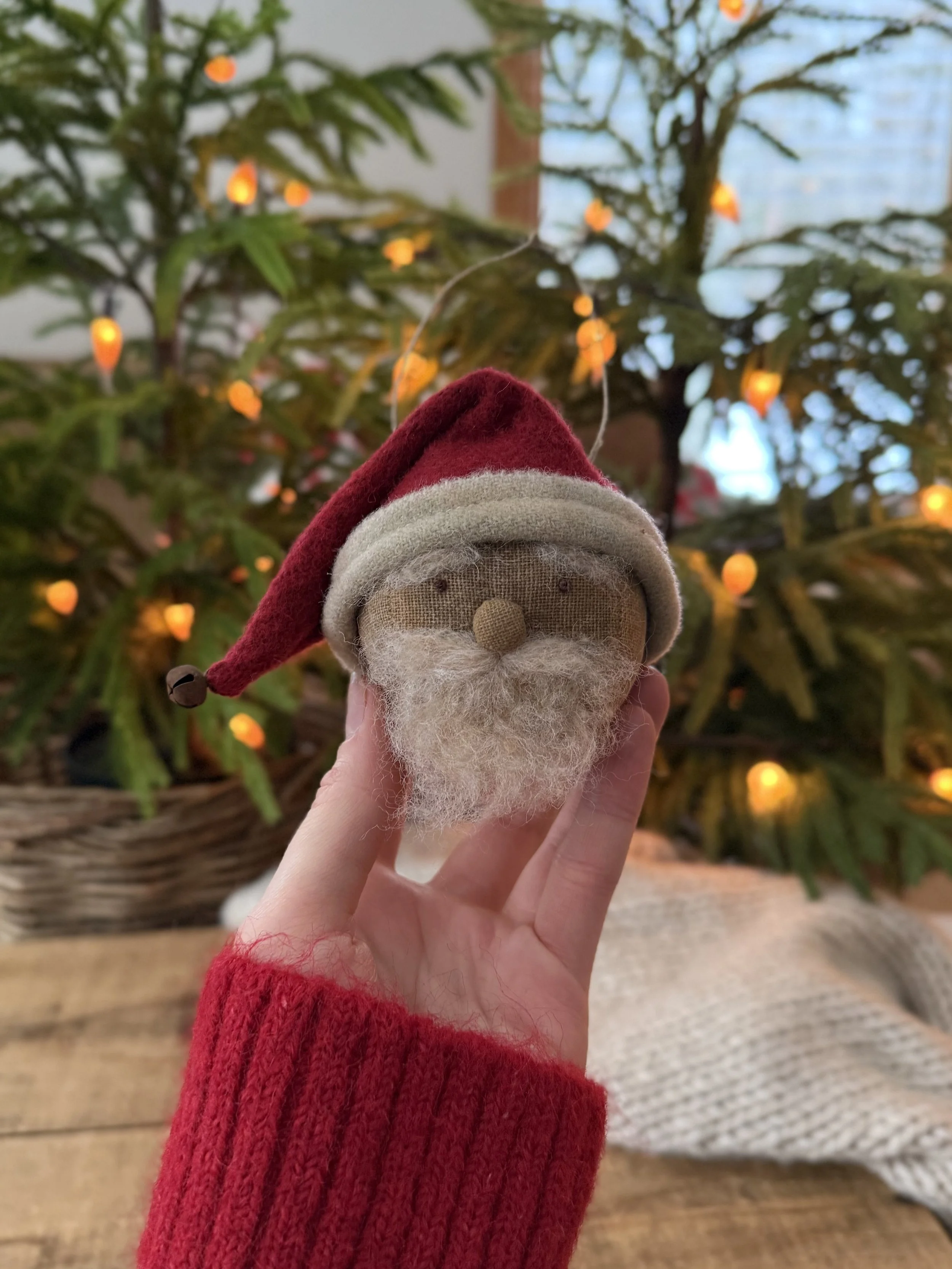 Felt Santa Claus ornament with red hat and white beard, held in front of decorated Christmas tree with yellow and orange lights.