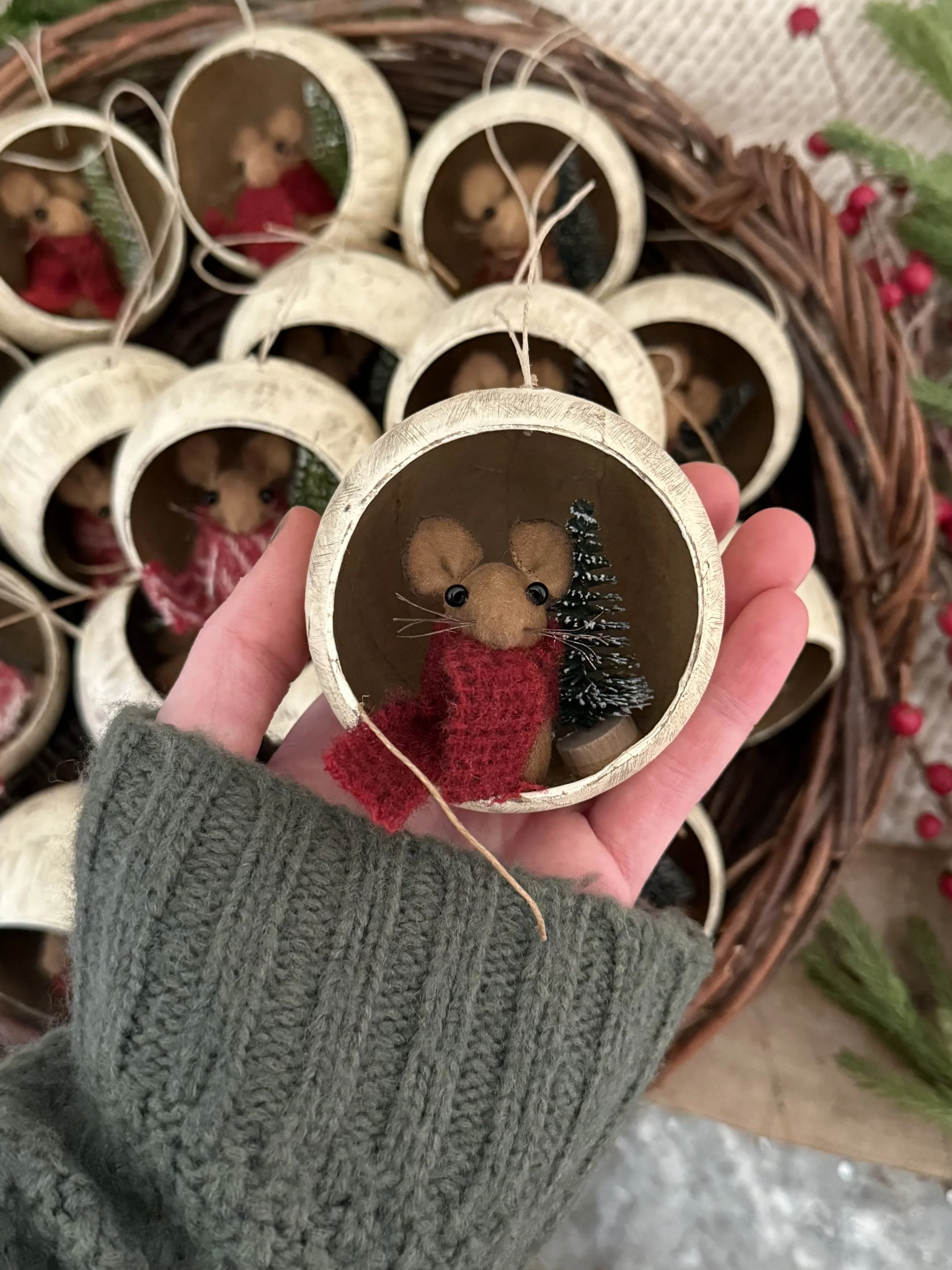 A person holding a small Christmas ornament featuring a felt mouse with black bead eyes, a red scarf, and a tiny green Christmas tree inside. Multiple similar ornaments are in a basket in the background.