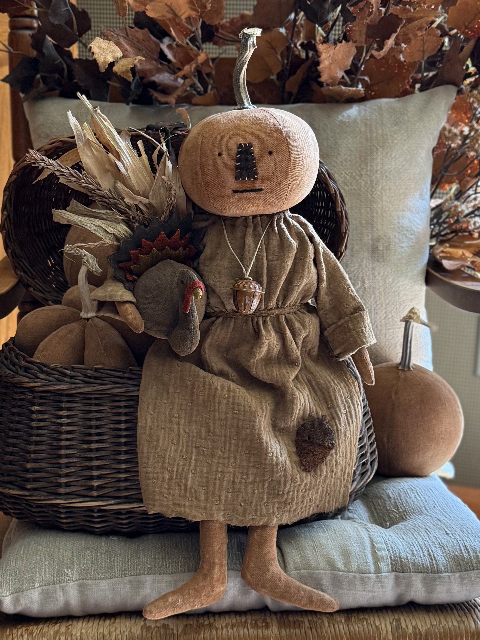 A fabric doll with a pumpkin head, sitting on a cushion, surrounded by autumnal decor including a basket of dried corn and pumpkins, and a background of fall foliage.