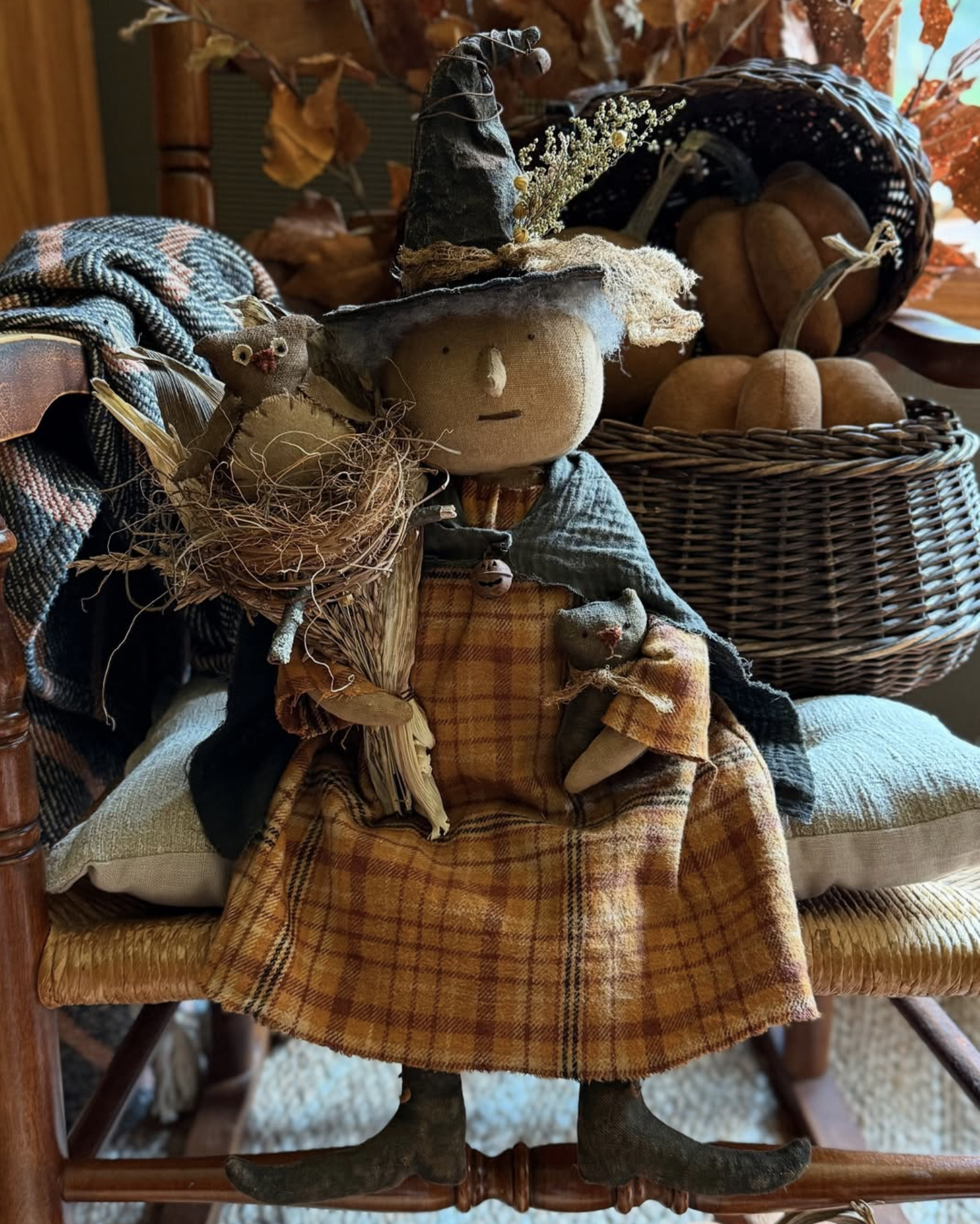 A rustic doll dressed as a witch with a pointy hat, sitting on a wooden chair with pillows. The doll holds a small broom and has a tiny stuffed animal. Behind, a basket of small pumpkin crafts and autumn leaves are visible.