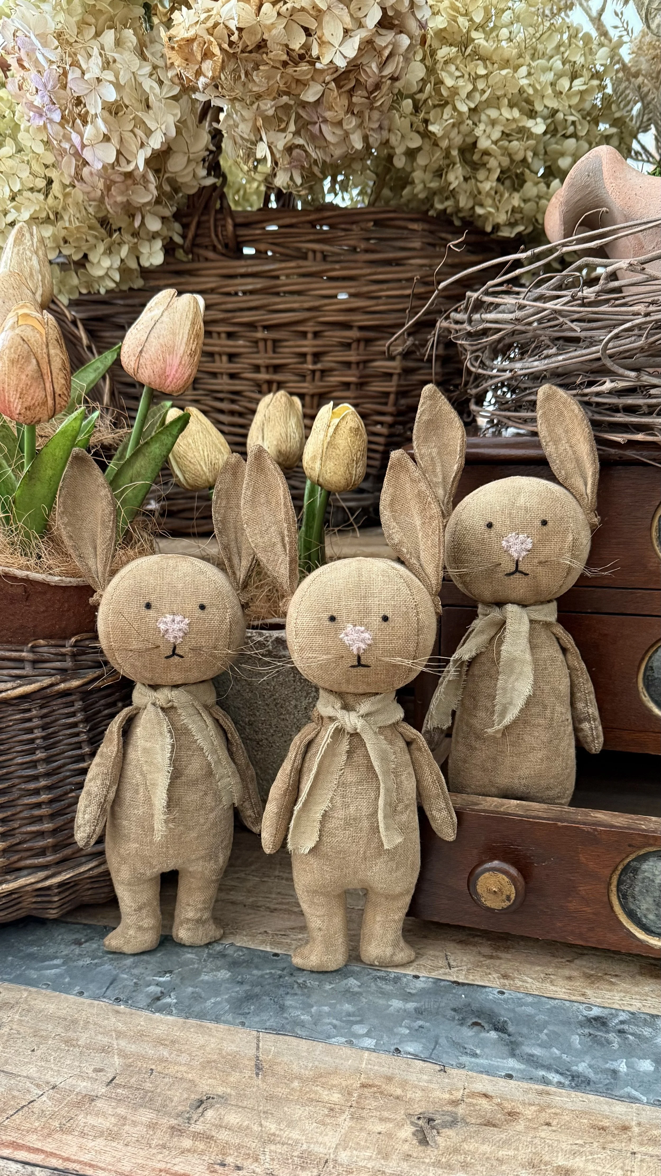 Three plush bunny toys with beige fabric bodies, long ears, and tied bows around their necks, standing in front of a basket of dried flowers and tulips, with wooden furniture and natural decor in the background.